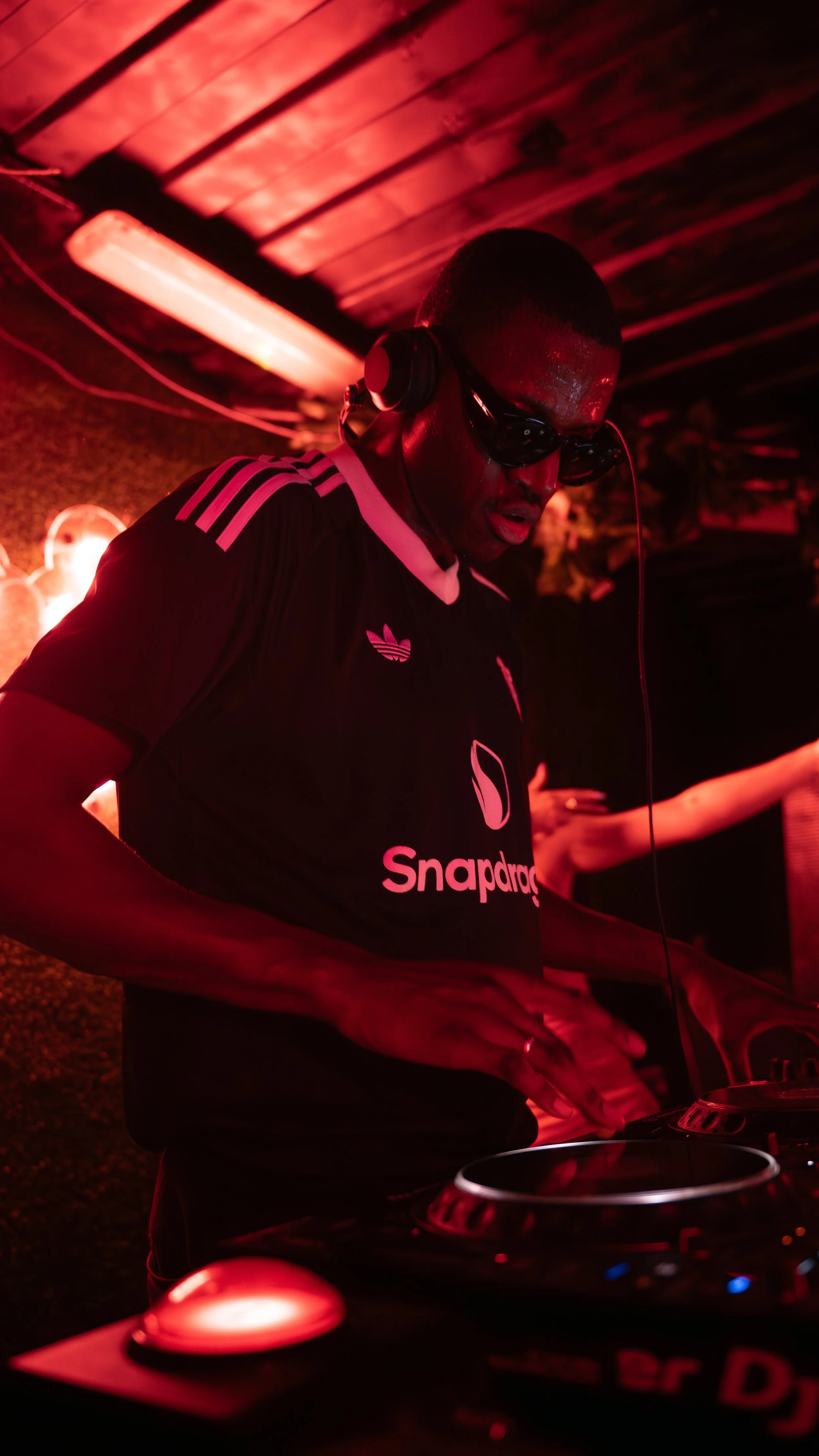 DJ wearing sunglasses and headphones operating a DJ controller in a dimly lit club with red lighting.