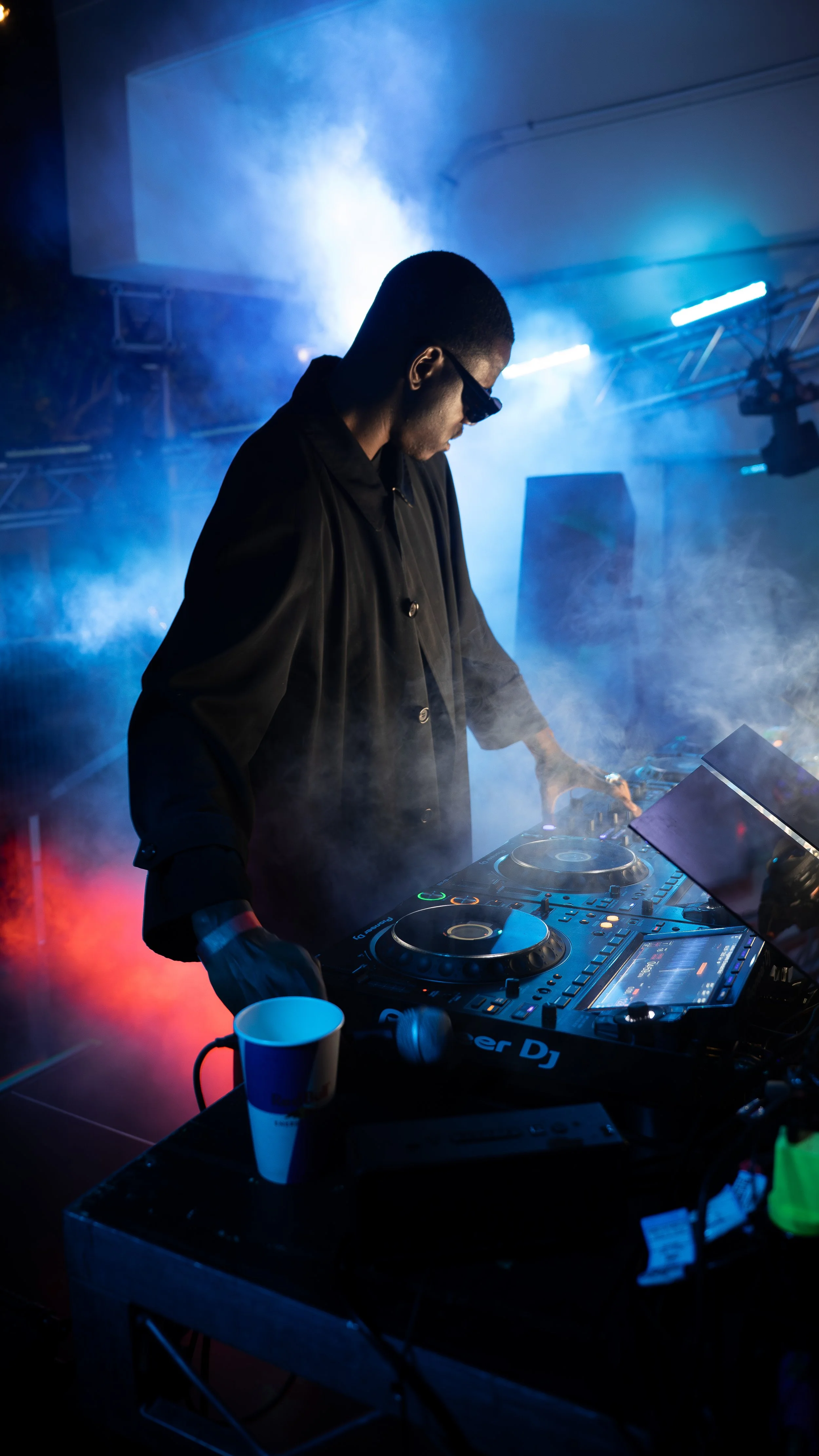 A DJ in dark clothing and sunglasses is performing at a nightclub with colorful lights and mist, working on a DJ mixing console.