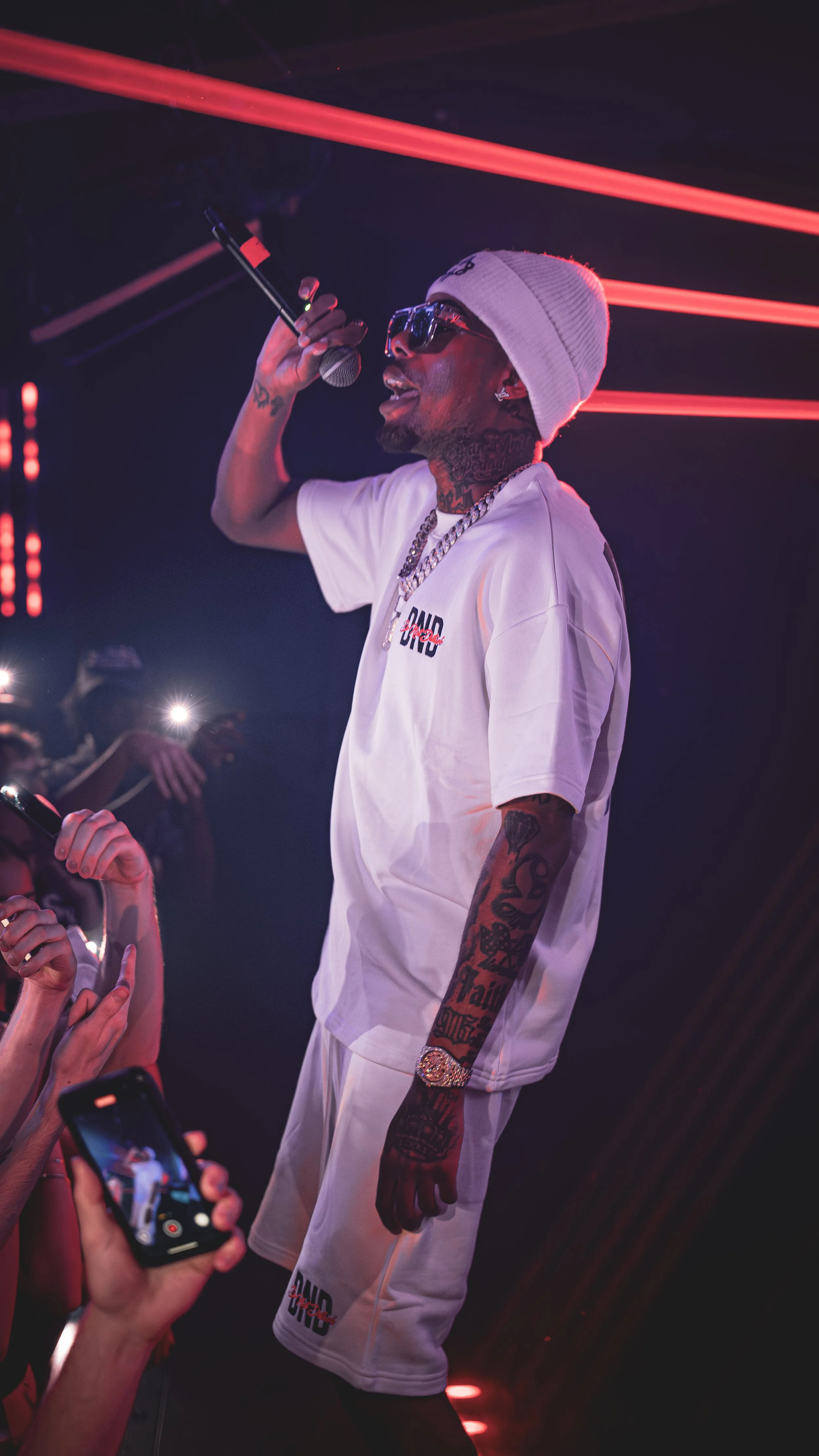 A male performer with tattoos, sunglasses, and a white beanie, holding a microphone on stage during a concert with red lighting.