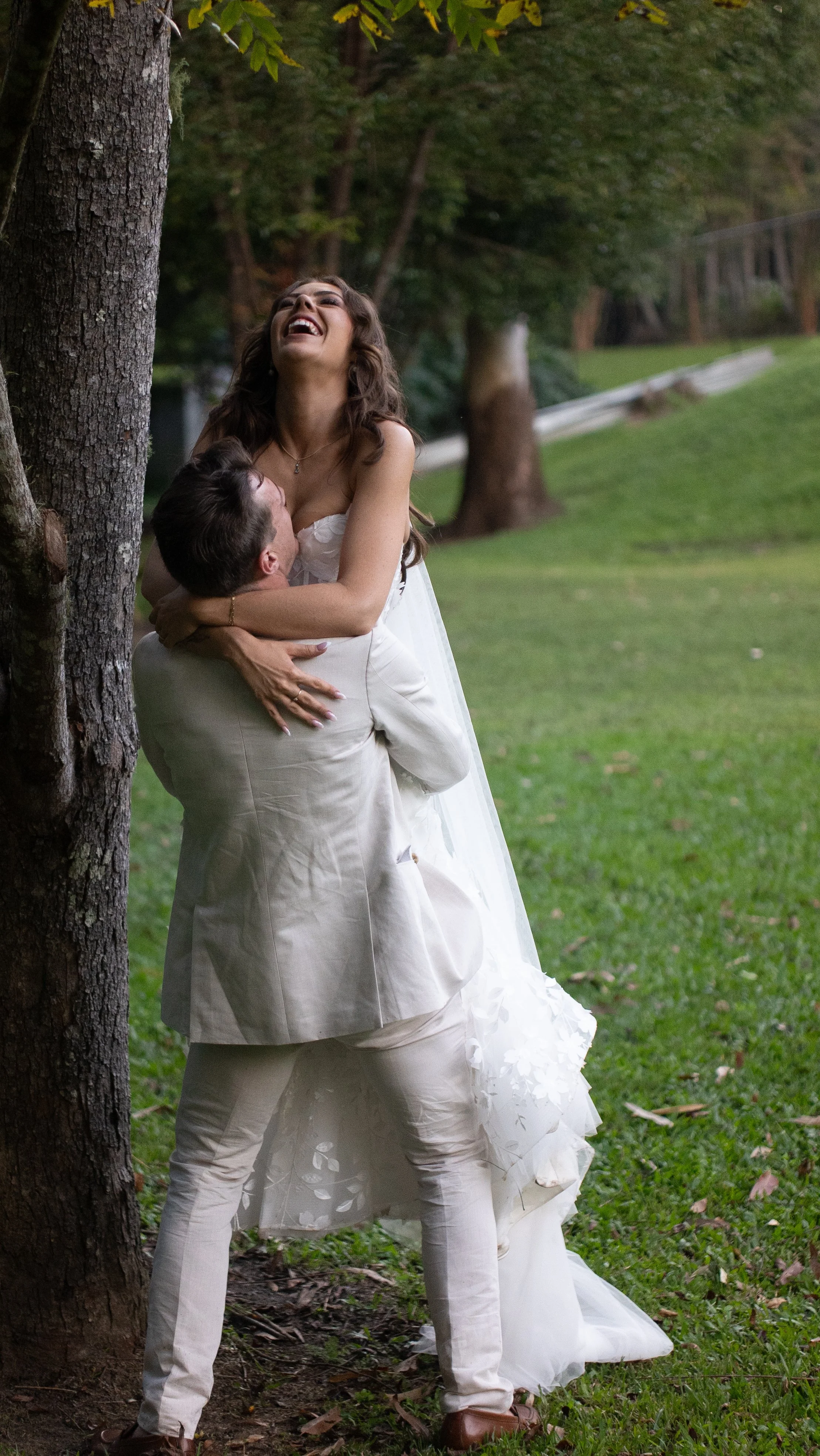 A couple celebrating outdoors, with the man lifting the woman who is dressed in a wedding gown against a tree, smiling and laughing.