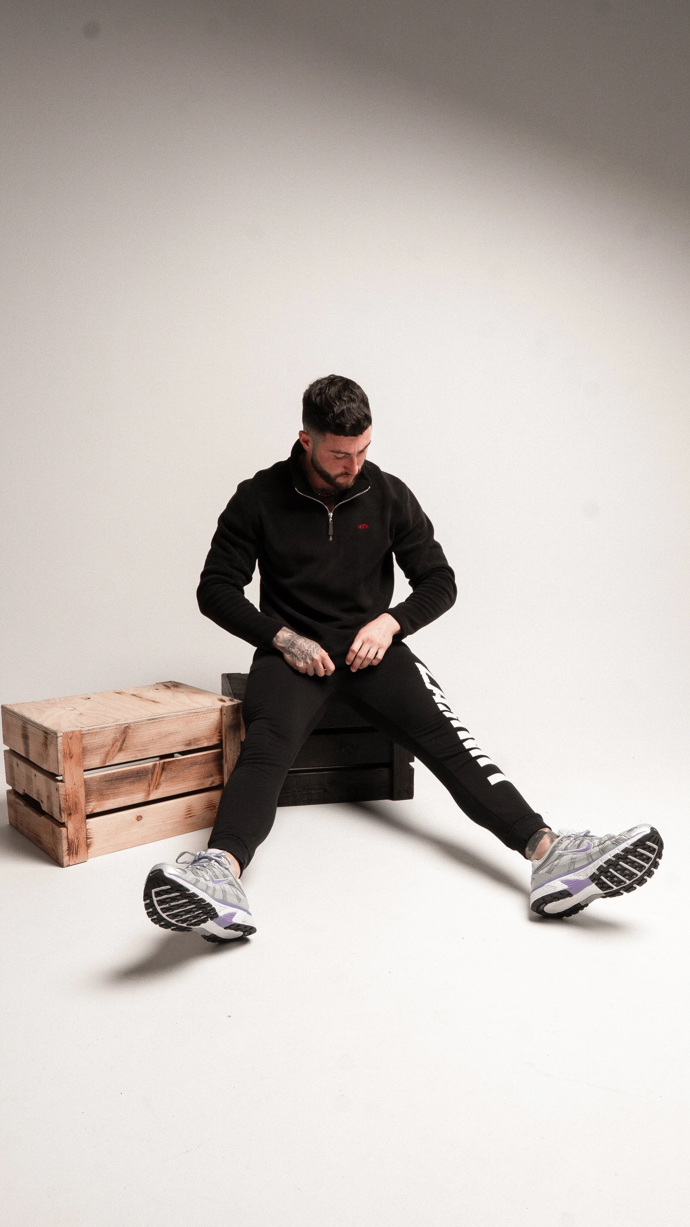 A man with tattoos on his arm sitting on a black box, stretching his legs apart while looking down, with a wooden crate beside him inside a plain white room.