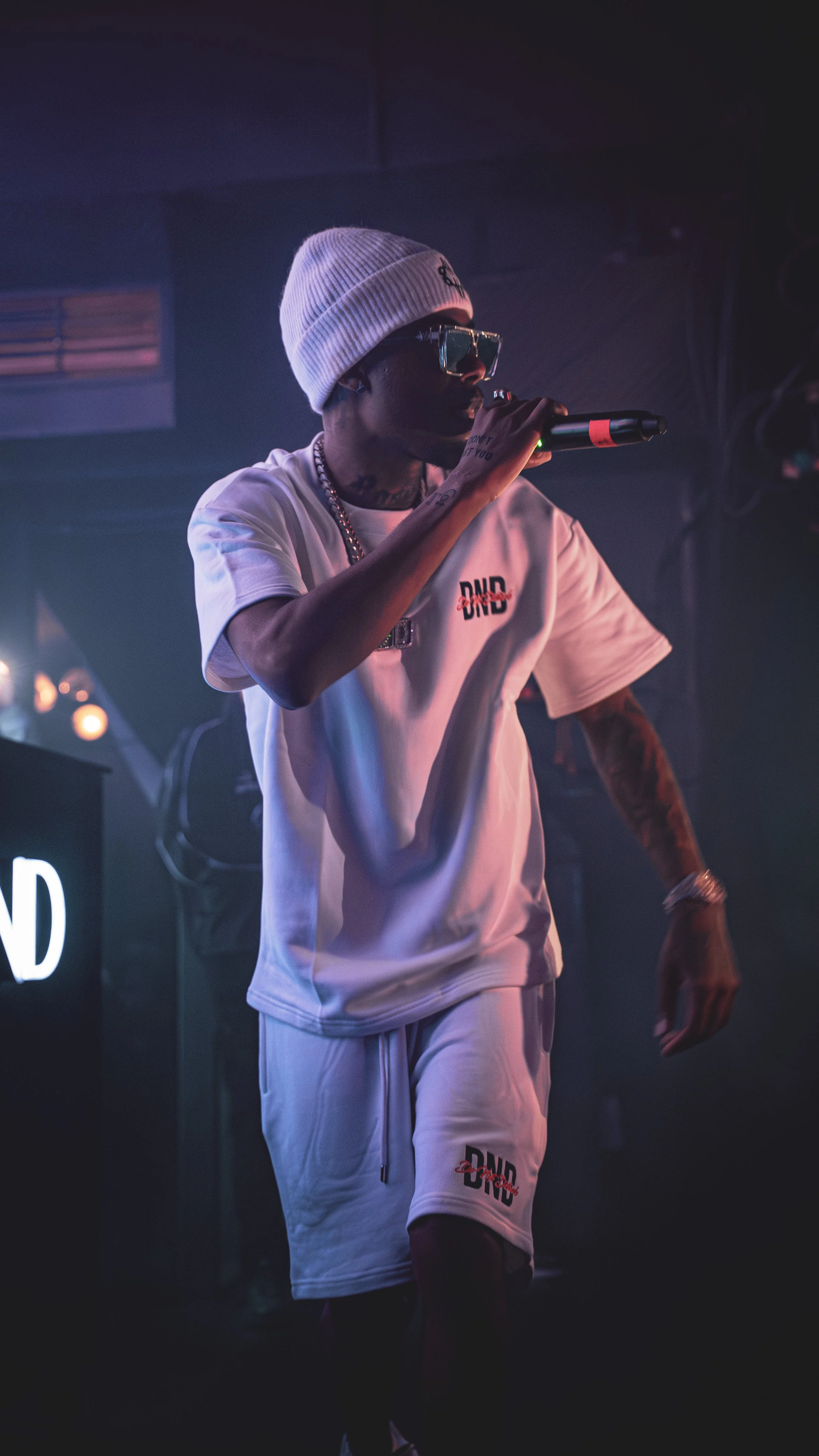 A man performing on stage, wearing a white beanie, sunglasses, a white t-shirt, and matching shorts, holding a microphone.