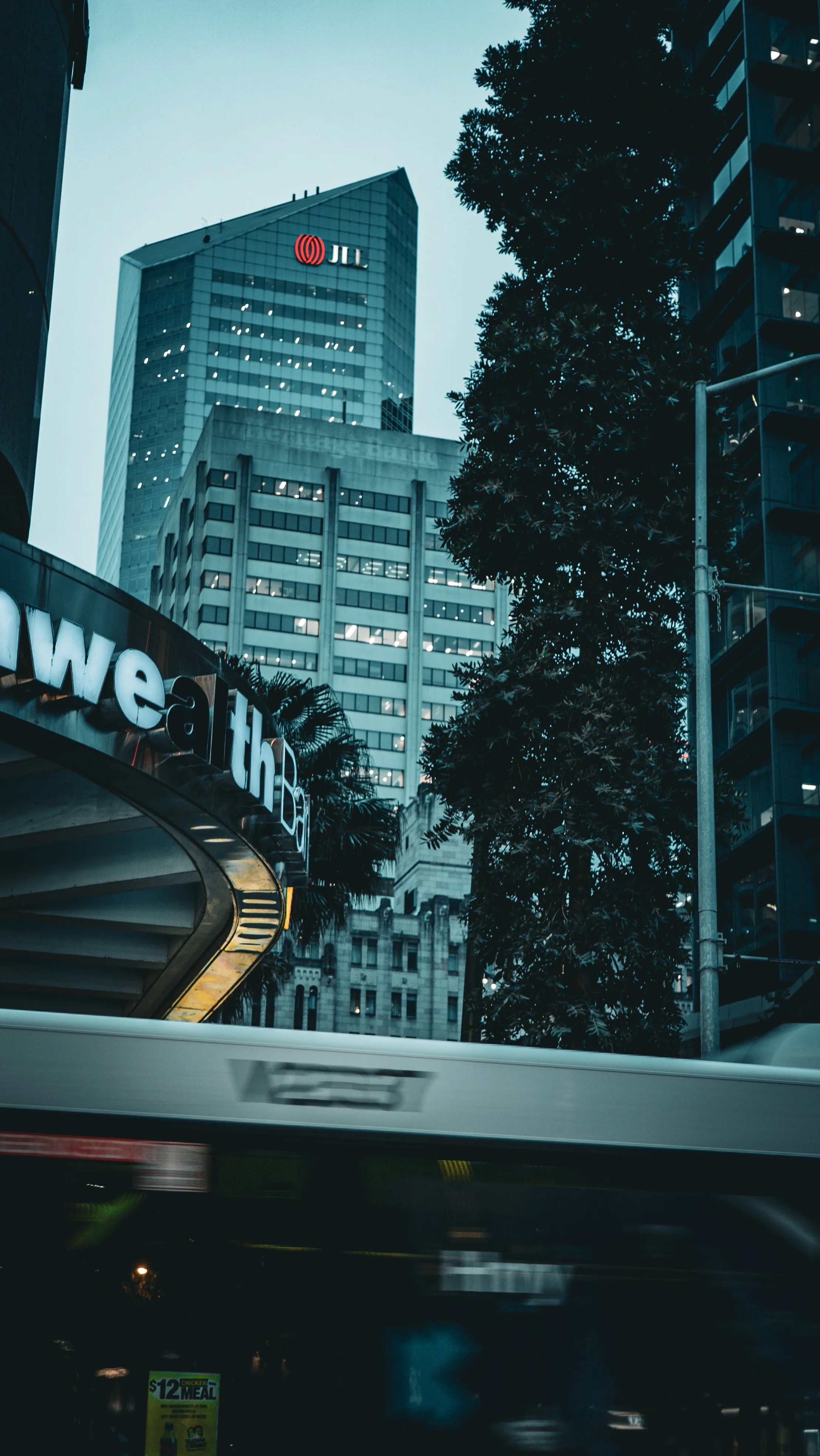 Cityscape with tall modern buildings, trees, a curved sign with the word 'Health', and a blurred moving vehicle in the foreground.
