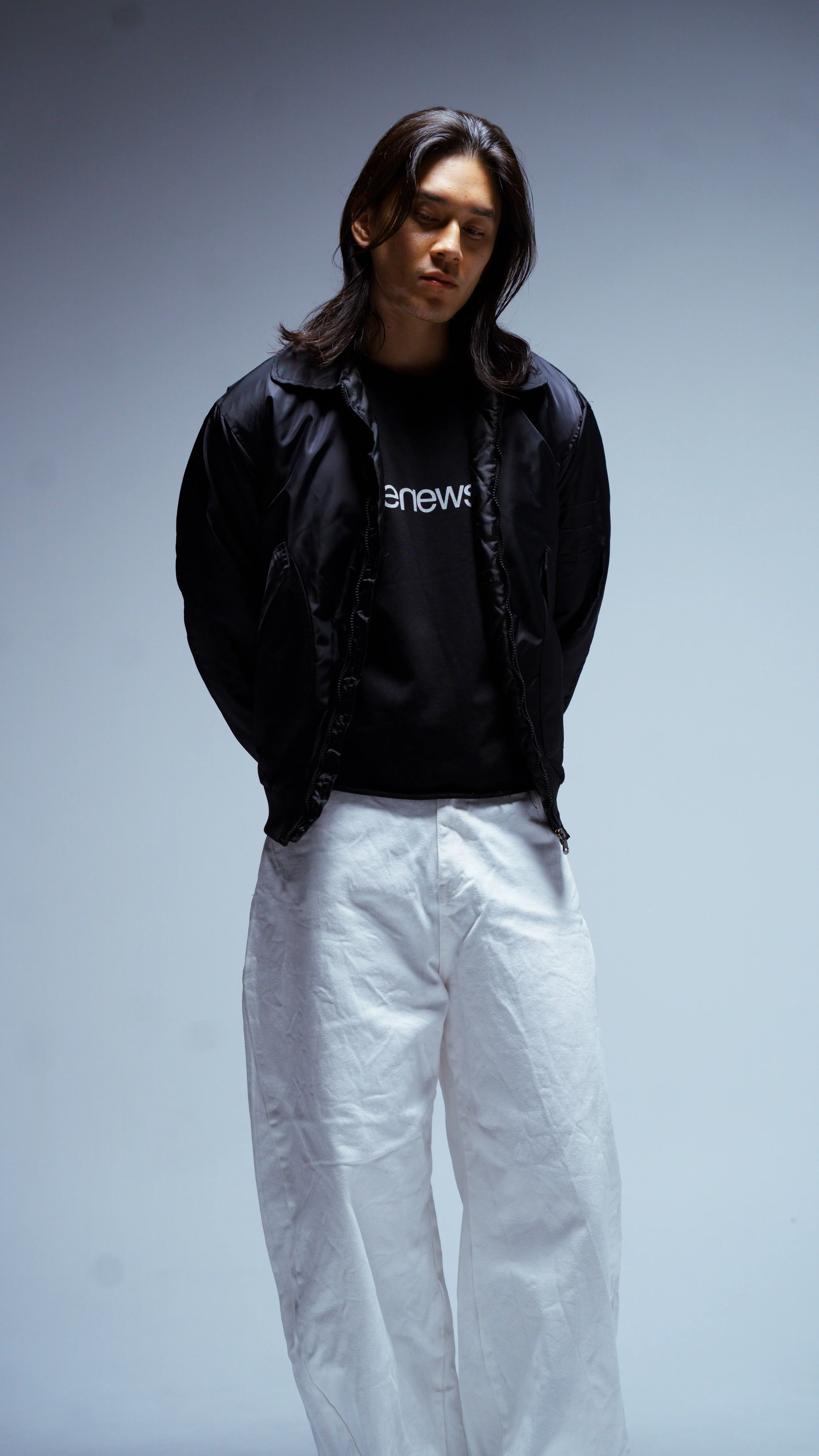 A young man with long dark hair wearing a black jacket, black shirt, and white pants, standing against a plain gray background.