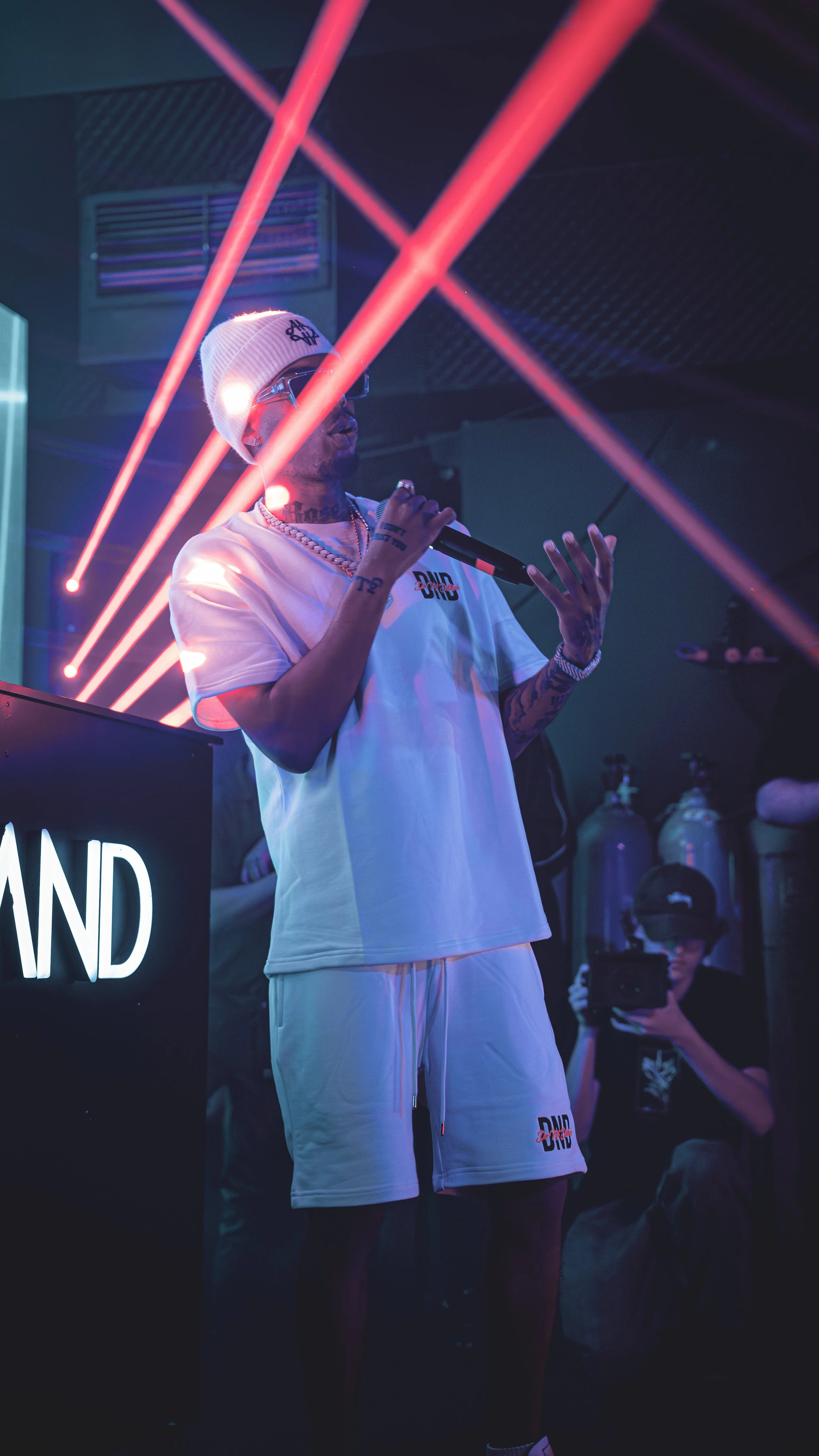 A man holding a microphone, wearing sunglasses, a beanie, and white shorts at a music event with stage lights and laser effects in the background.