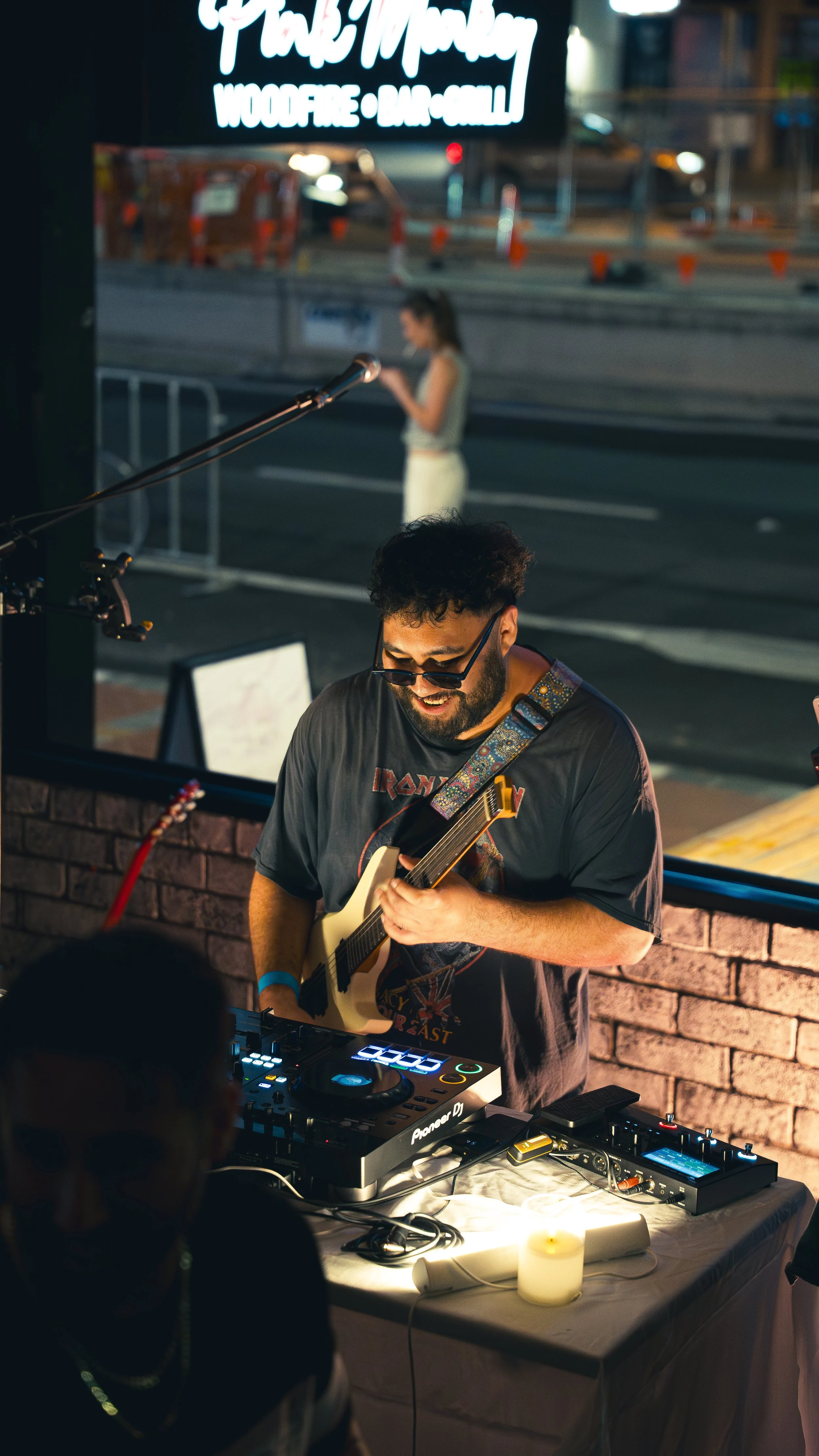 A male musician playing an acoustic guitar at a nighttime outdoor event, standing in front of DJ equipment on a table, with a candle beside him. Behind him, a woman is walking on the street, looking at her phone, with a bus stop and construction barr