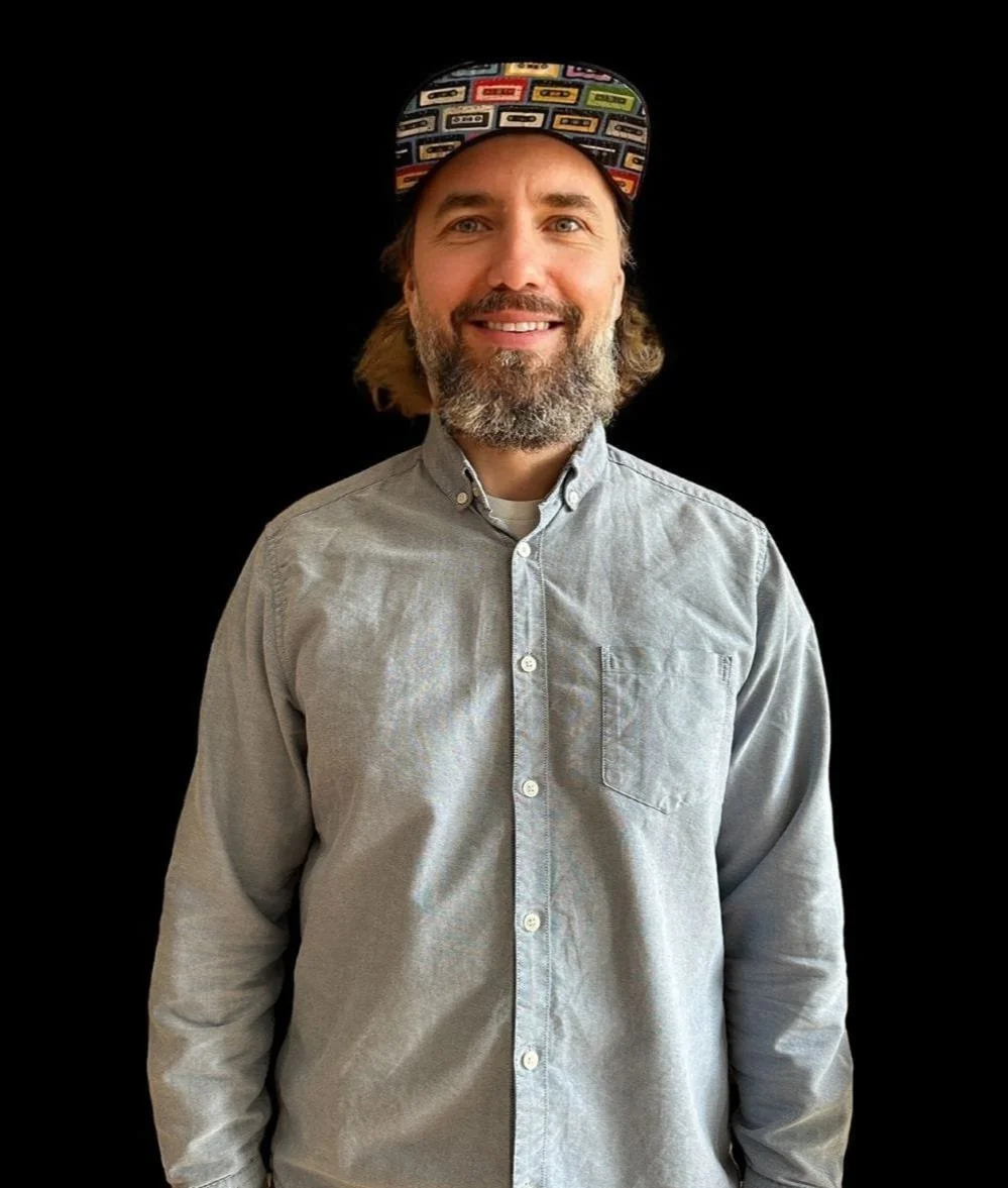 Ein lächelnder Mann mit einem grauen Hemd und eines bunten Basecaps vor einem schwarzen Hintergrund.