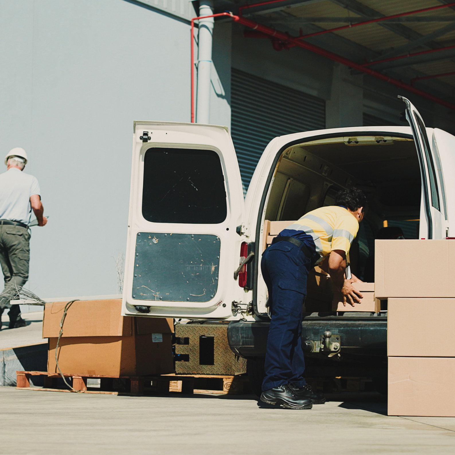 Workers loading cardboard boxes into the back of a white delivery van outside a building.