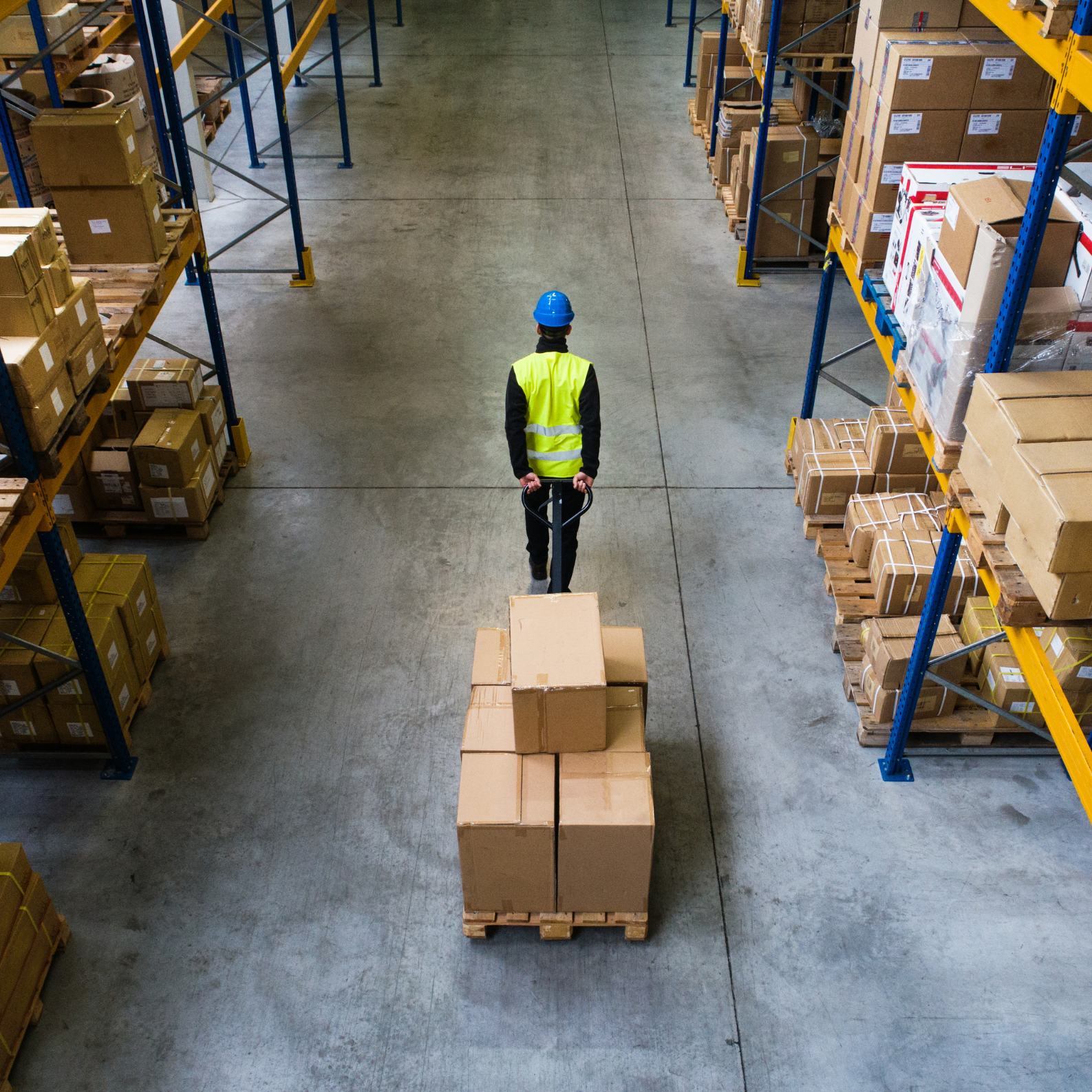 A worker in a warehouse wearing a blue hard hat and yellow reflective vest, pushing a pallet jack with boxes on a wooden pallet.
