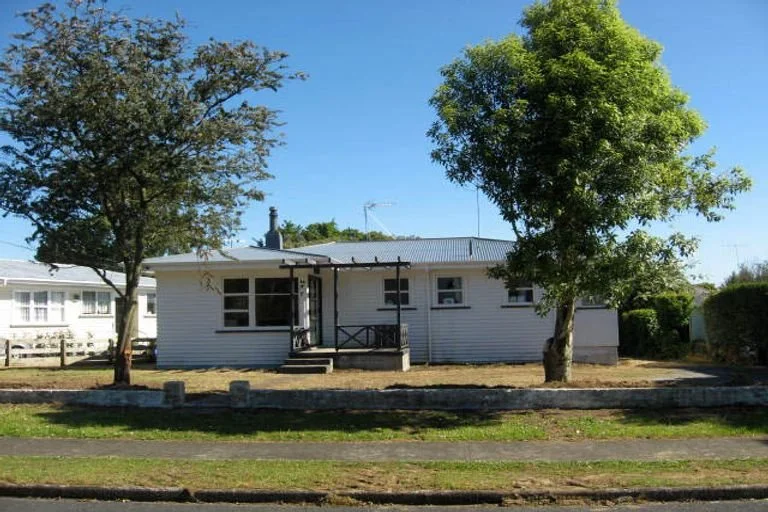 A small white house with a metal roof, two trees in front, and a small porch with steps. The grass in the yard appears dry, and there is a sidewalk and street in the foreground.