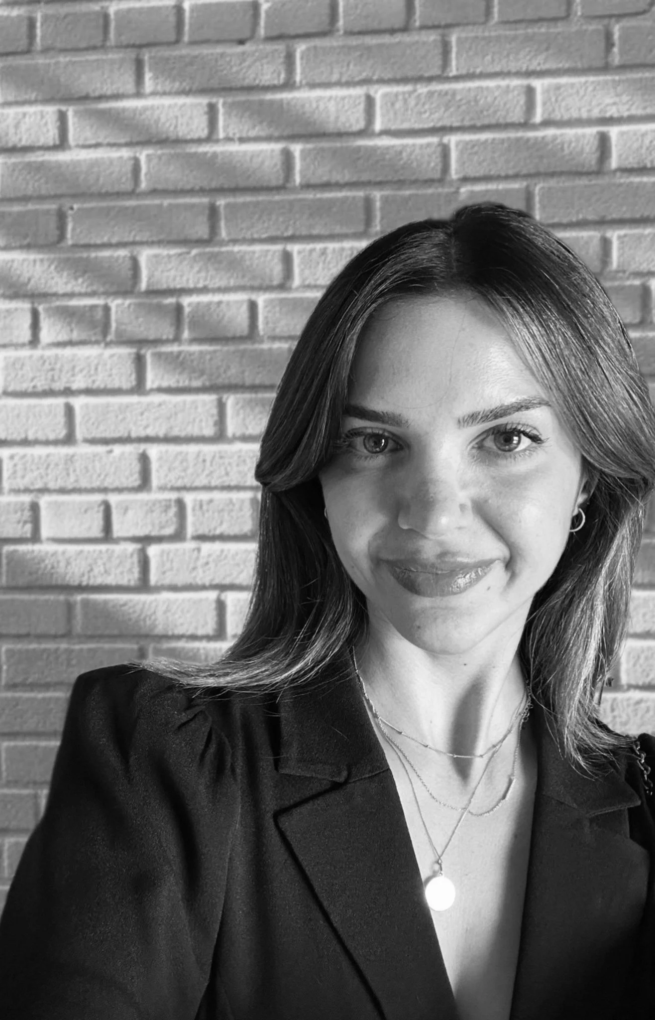 Black and white photo of a young woman with shoulder-length hair, wearing a blazer and layered necklaces, standing in front of a brick wall.