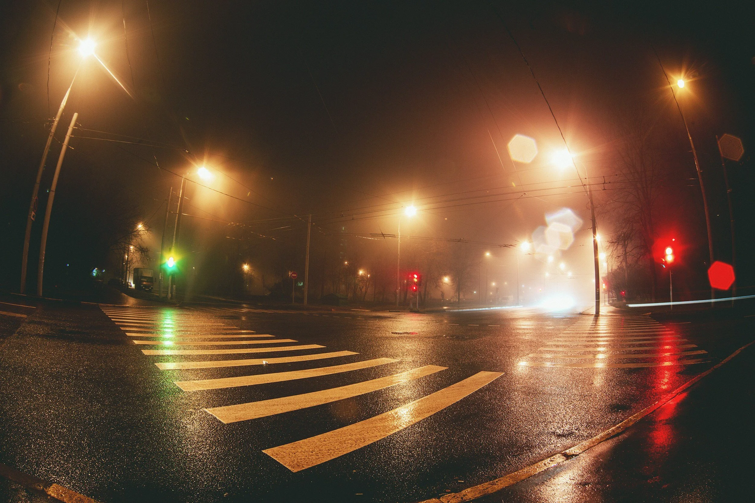 Quiet city street at night with wet pavement, streetlights, traffic signals, and light reflections, with some fog and rain.