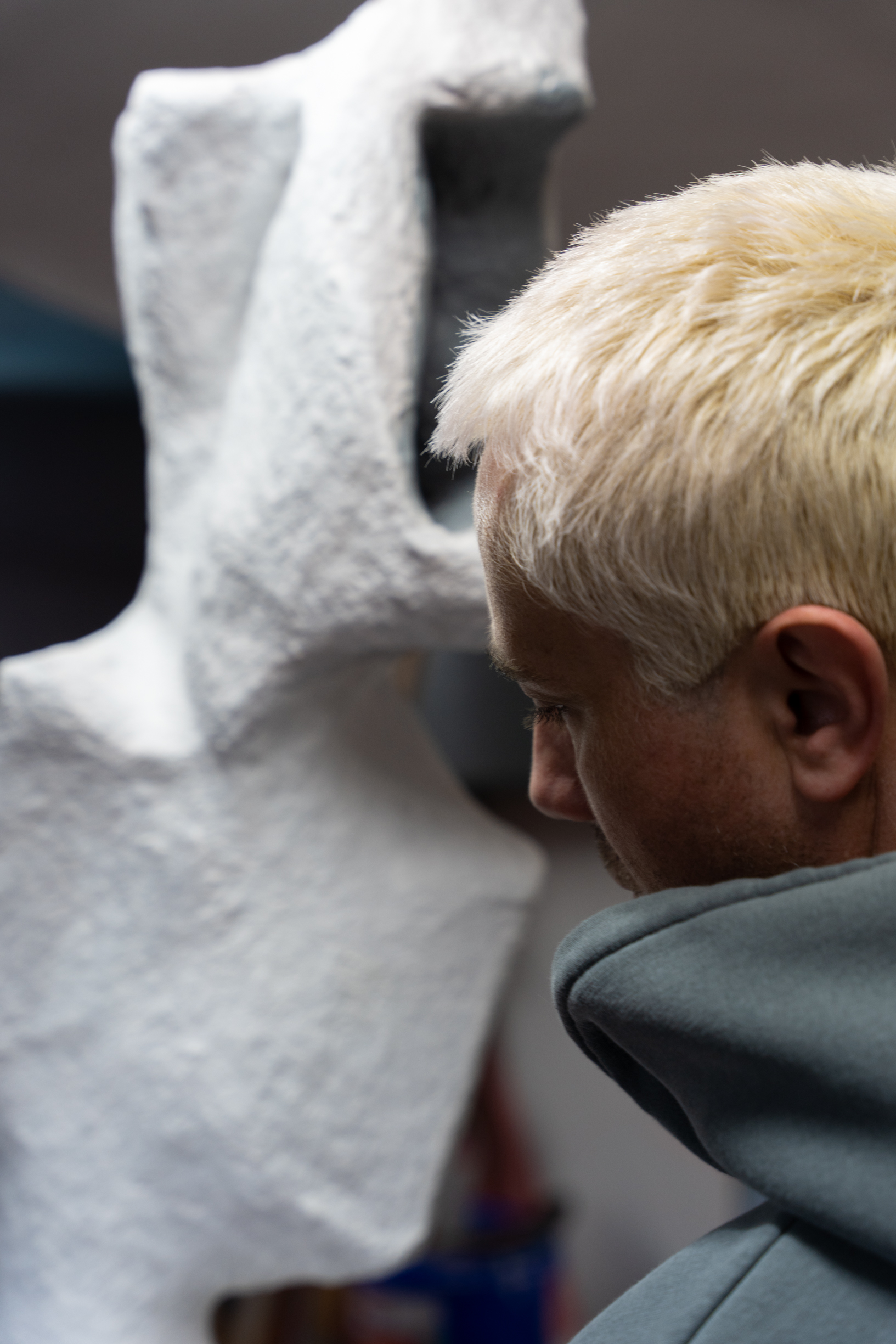 A person with blond hair and a gray hoodie is closely observing a large, white, textured sculpture of a face.