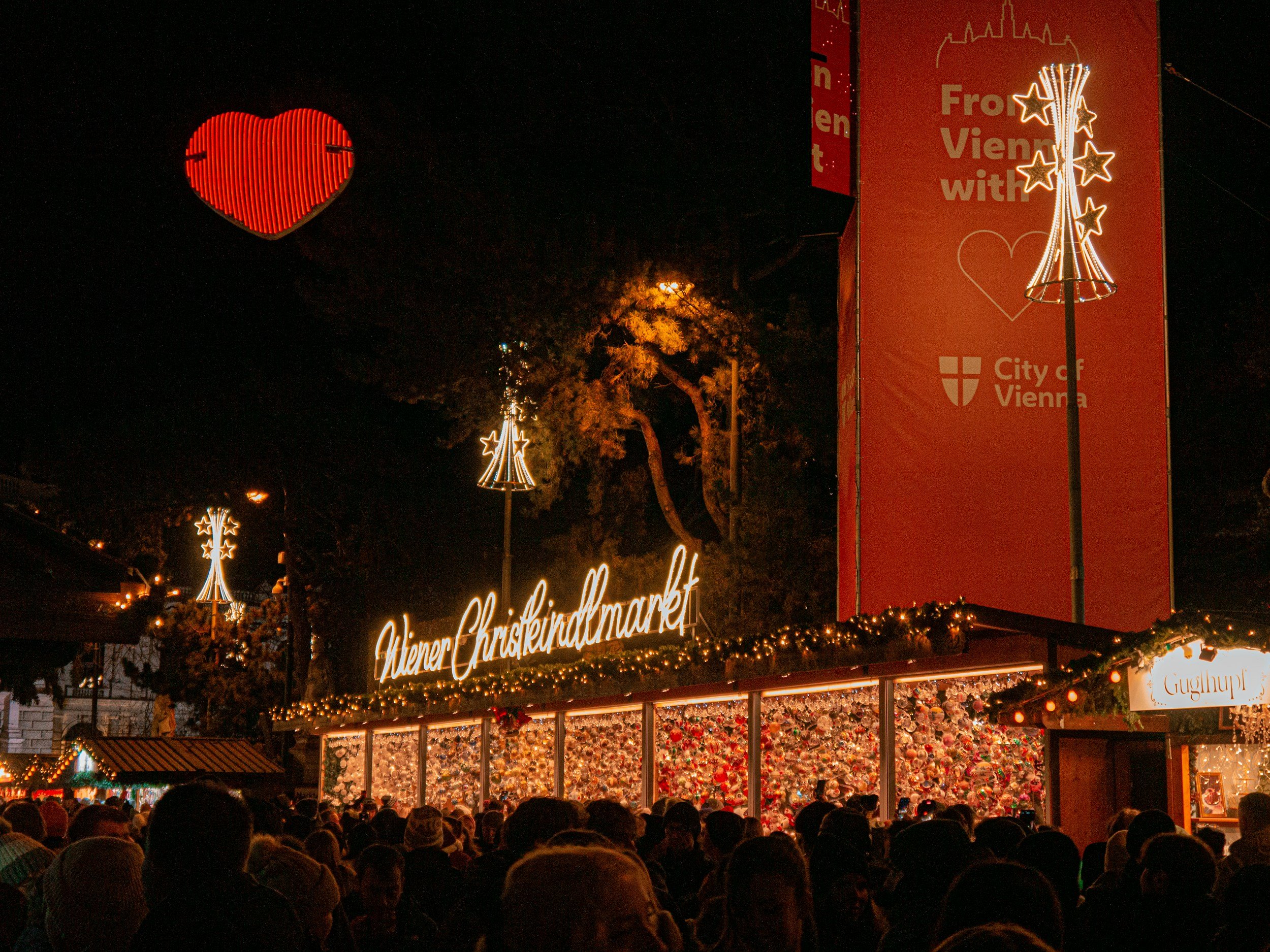 Night scene at Wiener Christkindlmarkt with festive lights, illuminated signs, and a crowd of people