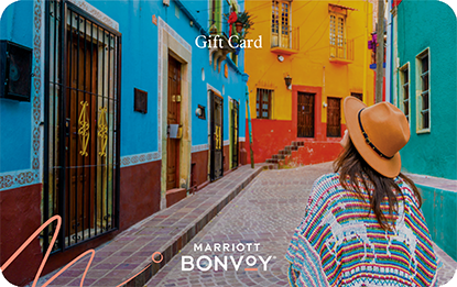 A woman with long hair wearing a hat and a colorful sweater walking through a vibrant, colorful street with blue, yellow, and orange buildings.