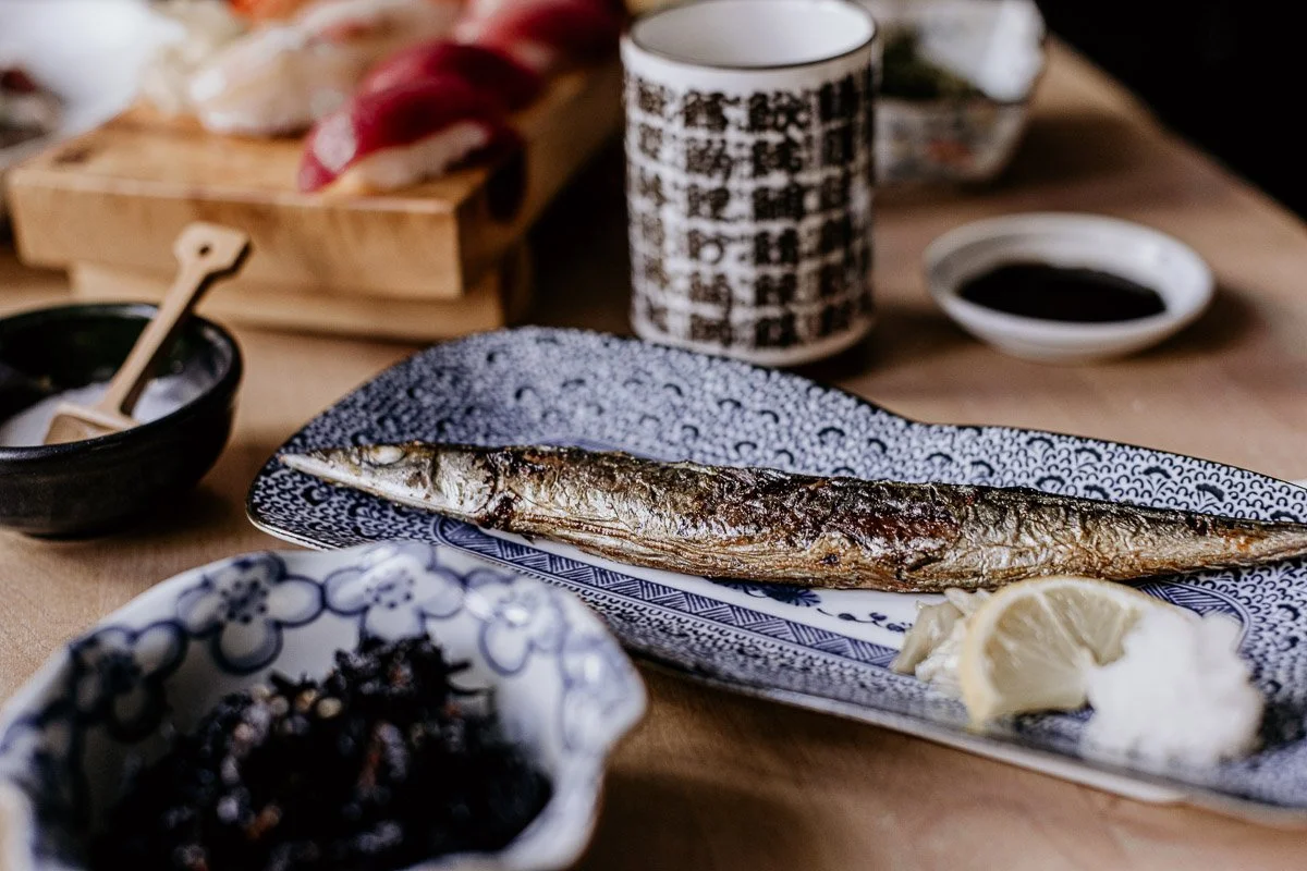 Gegrillter Fisch mit Zitronenscheiben, serviert auf einem blauen Geschirr, umgeben von Sojasauce, Japanischem Curry und weiteren Speisen auf einem Holztisch.