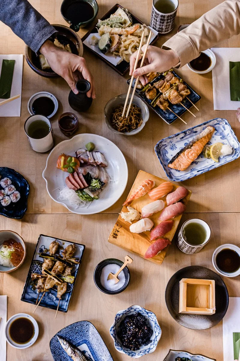 Verschiedene japanische Gerichte auf einem Tisch, darunter Sushi, Sashimi, Yakitori, Tempura, und Sojasauce, mit Tee und Geschirr.