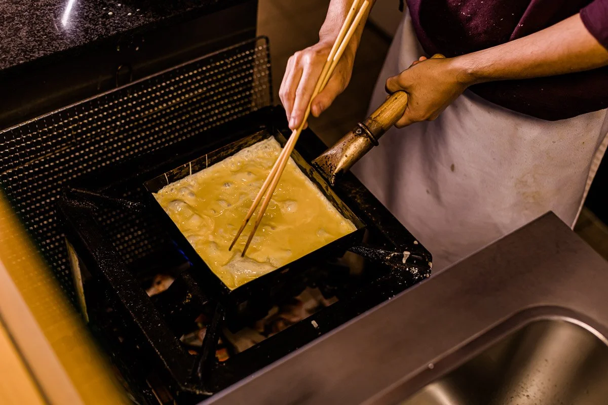 Das Bild zeigt eine Person, die Tofu in einer Pfanne frittiert, indem sie ihn mit Essstäbchen dreht, während sie die Pfanne mit einer Zange hält.
