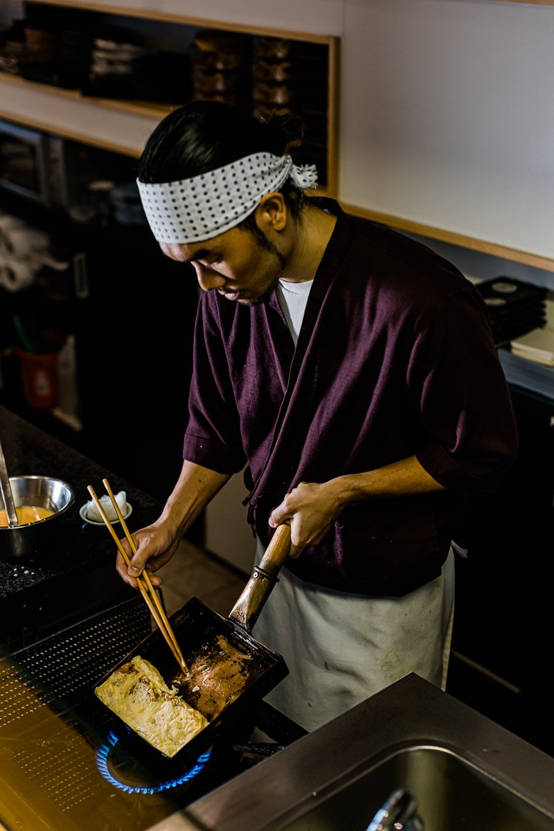 Ein Koch bereitet Eier in einer Pfanne auf einem offenen Gasherd zu, während er bückt und mit Essstäbchen arbeitet.
