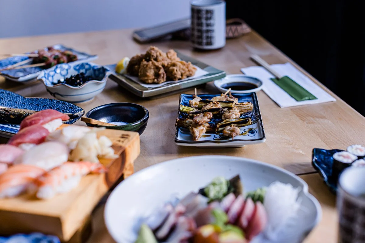 Verschiedene japanische Sushi- und Sashimi-Gerichte auf Tellern und Tabletts, umgeben von Sojasauce, Sushi-Rollen und anderen Beilagen auf einem Holztisch.