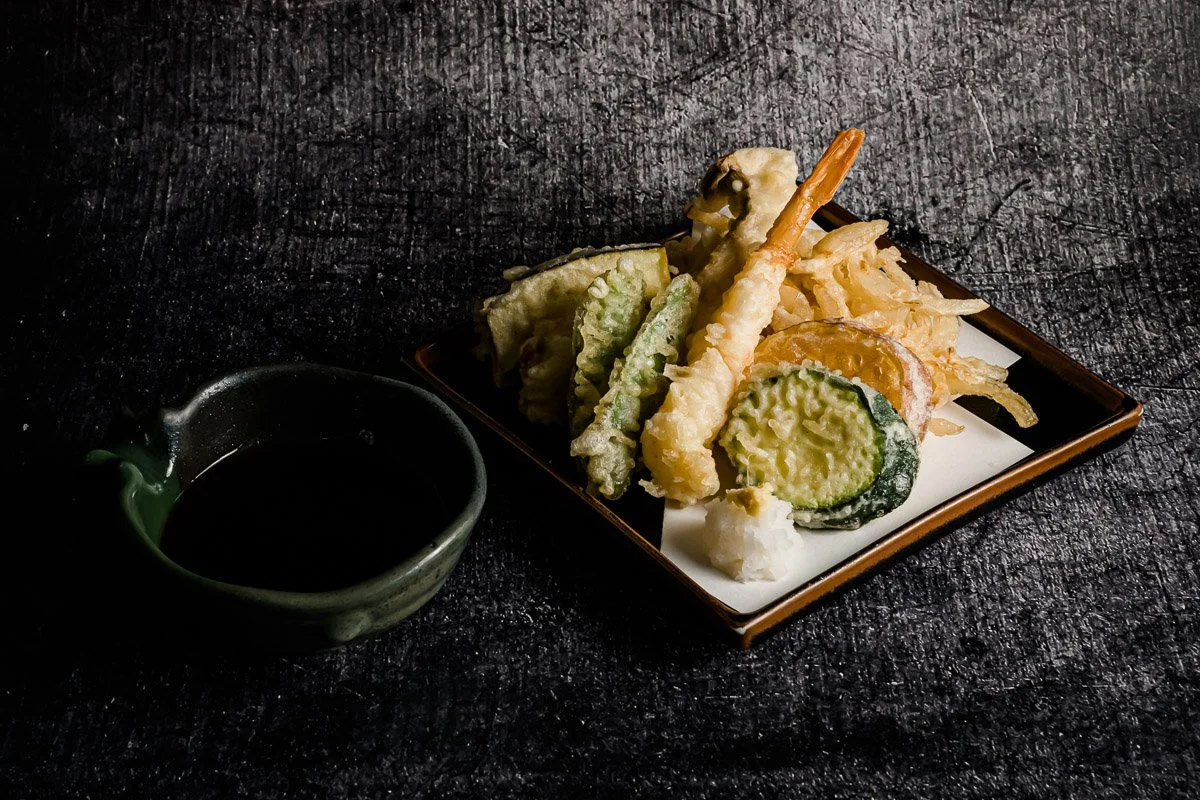 Tempura-Gemüse auf einem schwarzen Teller mit Sojasoße in einer kleinen Schale auf dunklem Hintergrund.