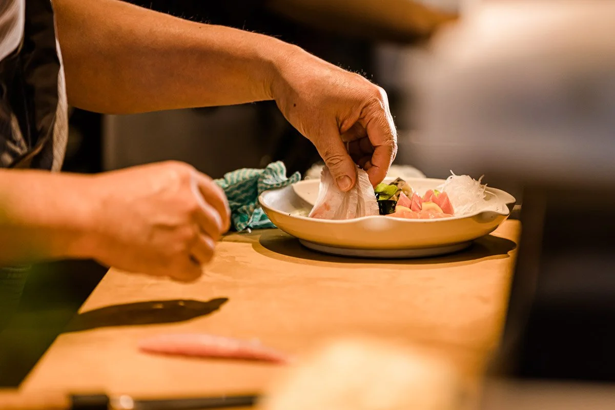Nahaufnahme einer Person beim Zubereiten von Sushi, die Fisch auf einen Teller legt.