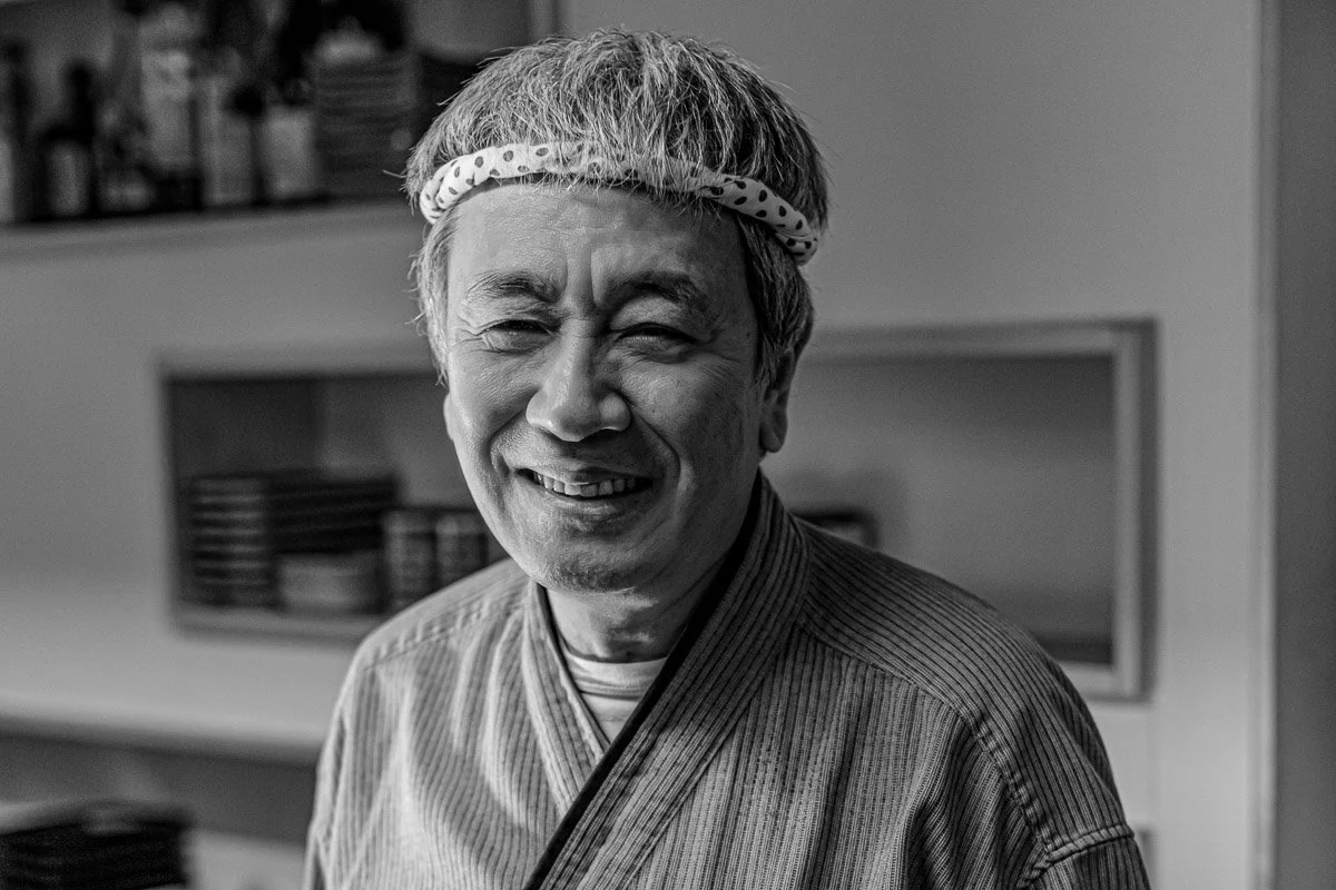 Ein lächelnder älterer Mann trägt ein Stirnband mit Punkten und steht in einer Küche, im Hintergrund befinden sich Schränke und Geschirr.