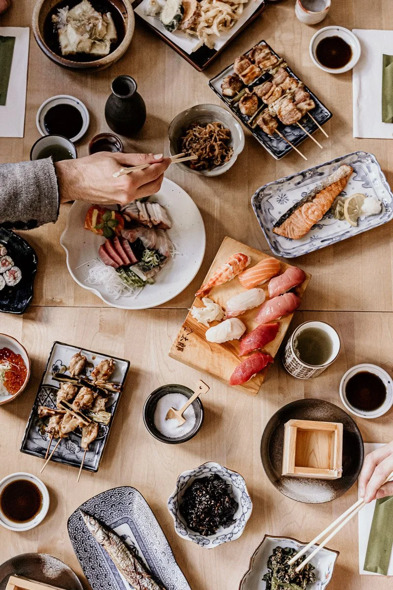 Ein japanisches Essen Tisches mit Sushi, Sashimi, gegrilltem Fisch, Suppen, Sojasauce, Tee und verschiedenen Beilagen, alles auf einer Holztischplatte.