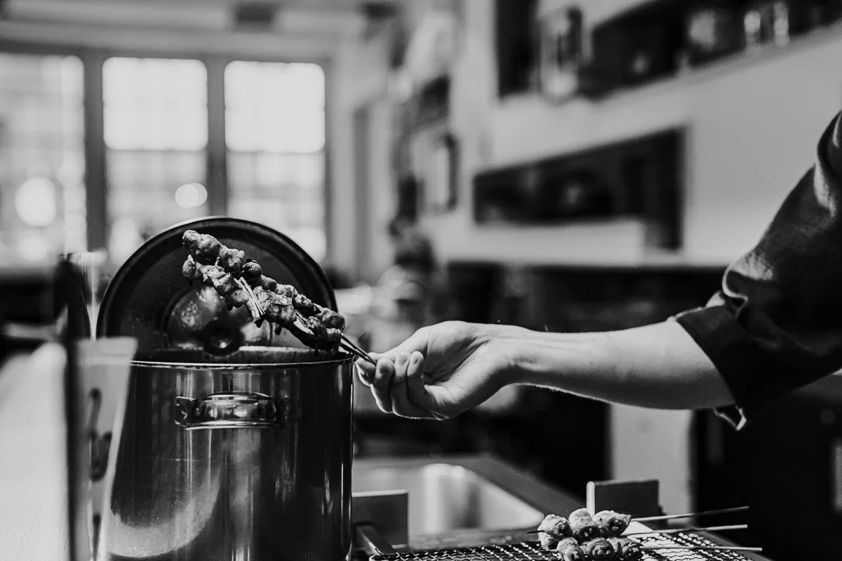 Eine schwarze und weiße Aufnahme zeigt eine Person, die mit einer Zange Hähnchenspieße aus einem großen Topf entnimmt, im Hintergrund sind Fenster mit Lichtdurchlässigen Vorhängen sichtbar.
