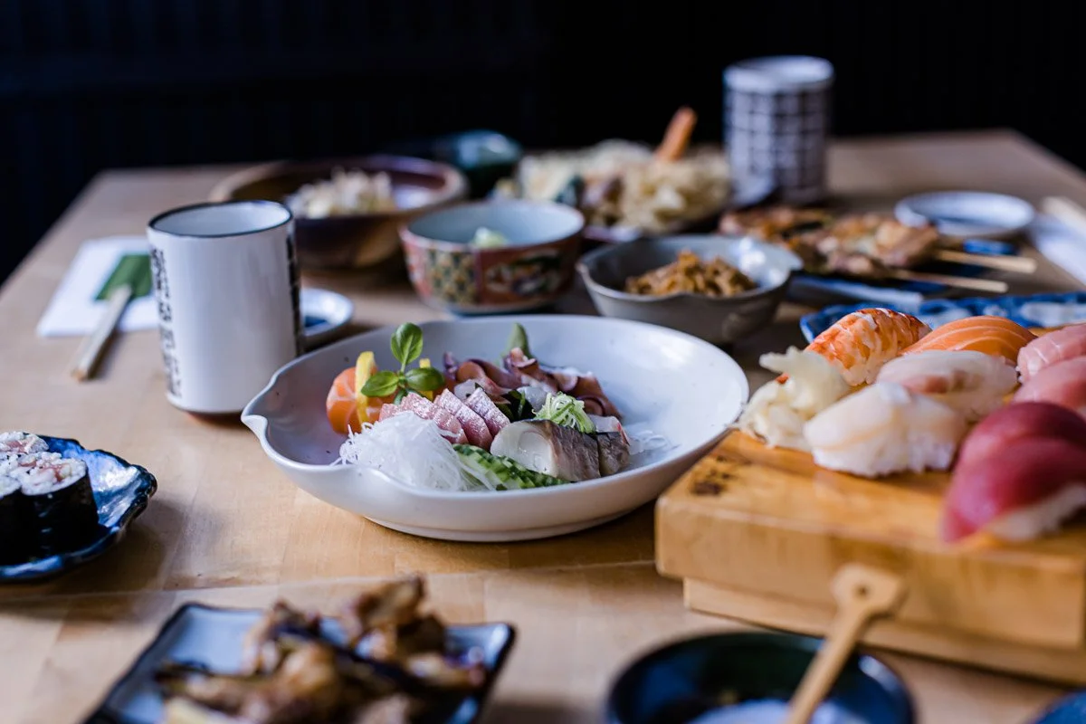 Verschiedene Sushi und sashimi auf einem Holztisch, serviert in Schalen und auf Holzbrett, dazu Getränke und Beilagen.