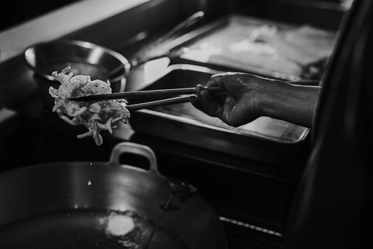 Hand hält Nudeln mit Essstäbchen über einer Pfanne, in der Öl brutzelt, Küche im Hintergrund.