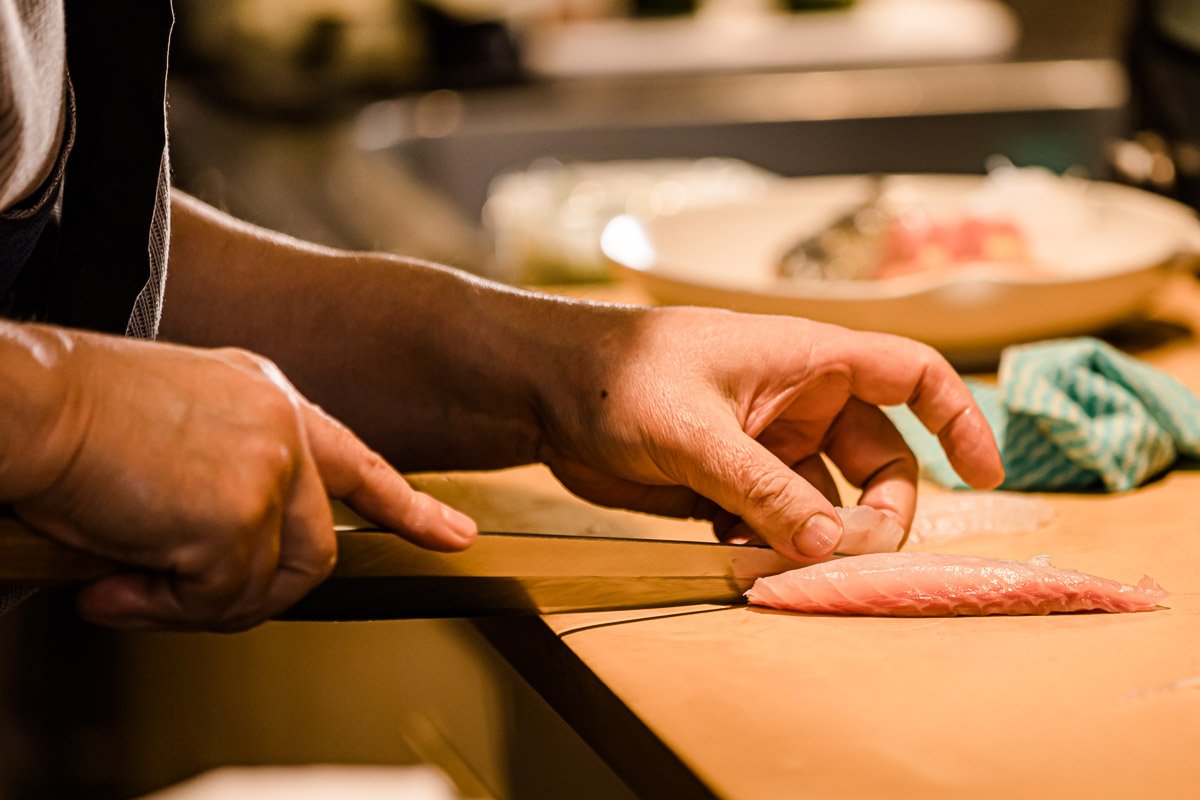 Person schneidet ein Stück rohes Fischfilet auf einem Schneidebrett in einer Küche.