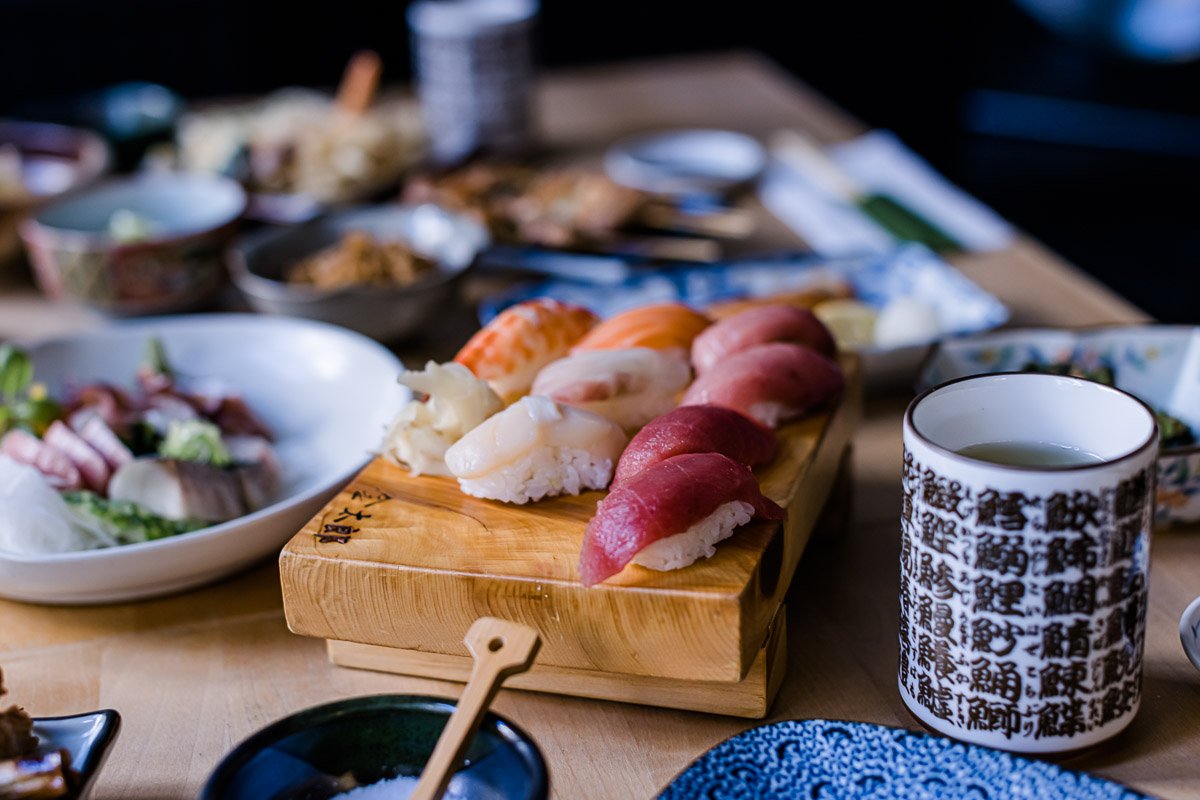 Verschiedene Sushi auf einem Holzbrett, daneben eine Tasse mit Tee und weitere Schüsseln mit japanischen Speisen auf einem Tisch.