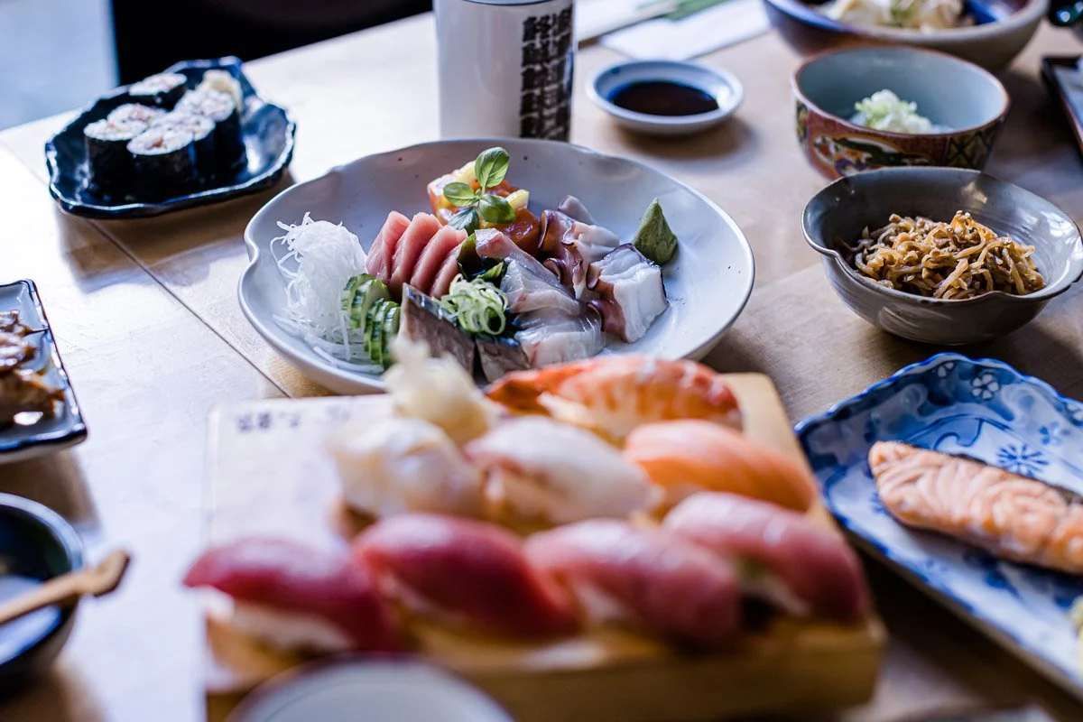 Verschiedene japanische Gerichte, darunter Sushi, Sashimi, Maki-Rollen, Nudeln und Lachsfilets, auf einem Holztisch.