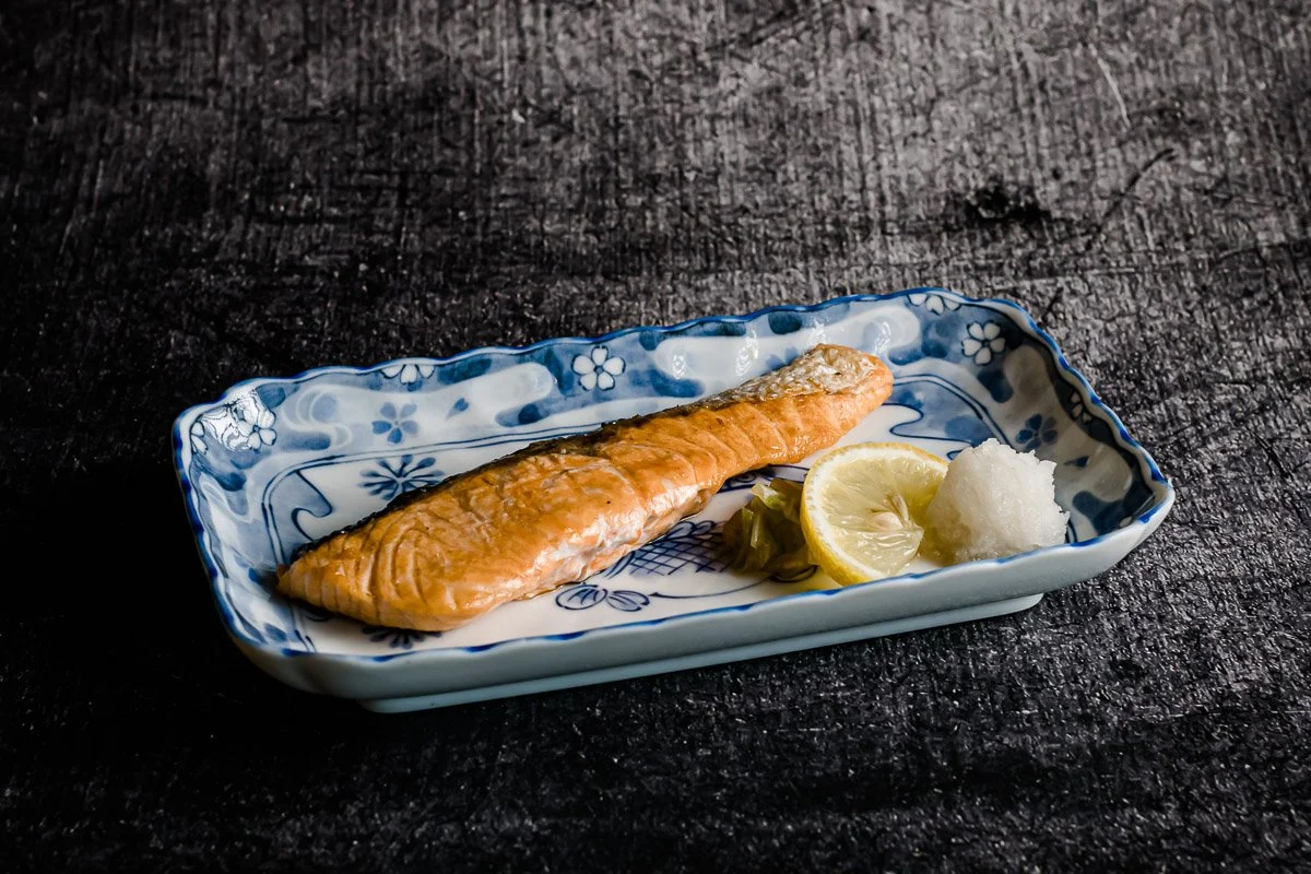 Gebratener Lachs mit Zitronenscheibe, Rettich und Salat auf einer blauen und weißen Keramikschale auf schwarzem Holztisch.