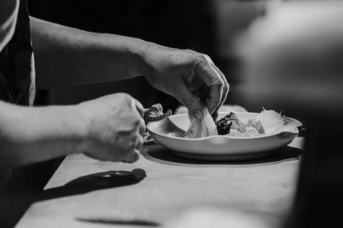 Eine Hand legt Essen auf einen Teller, vermutlich beim Servieren in einer Küche. Das Bild ist schwarz-weiß.