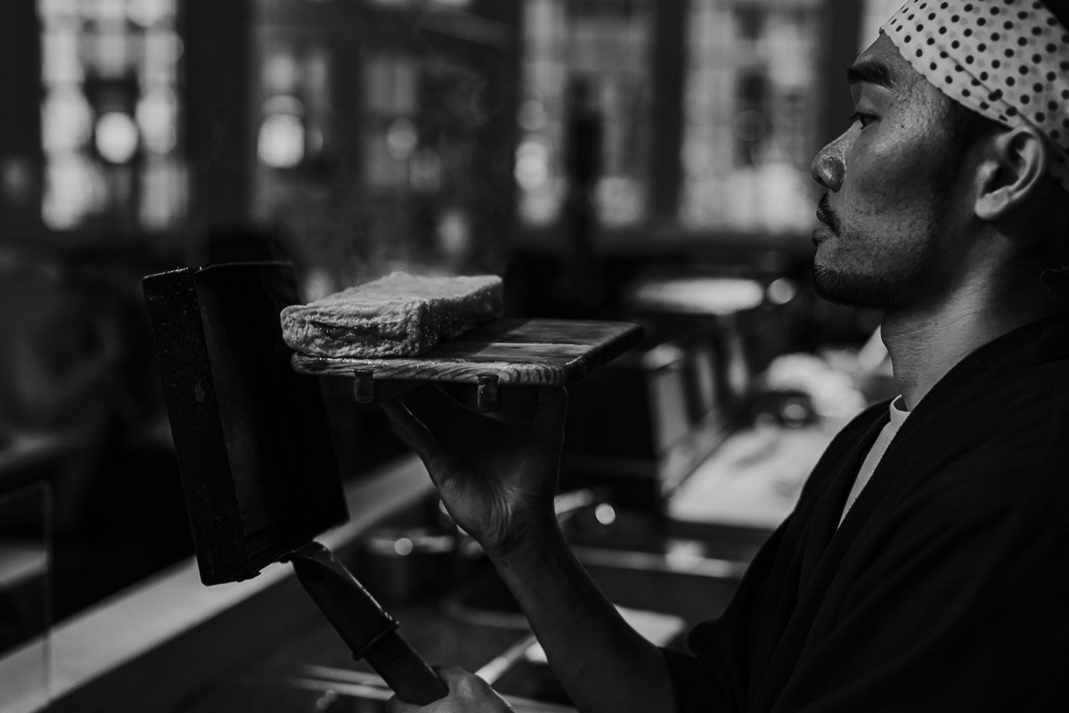 Ein Mann mit einem Kopftuch hält eine Holzplatte mit einem Stück Fleisch vor sich, während er in einem Restaurant oder Café sitzt.