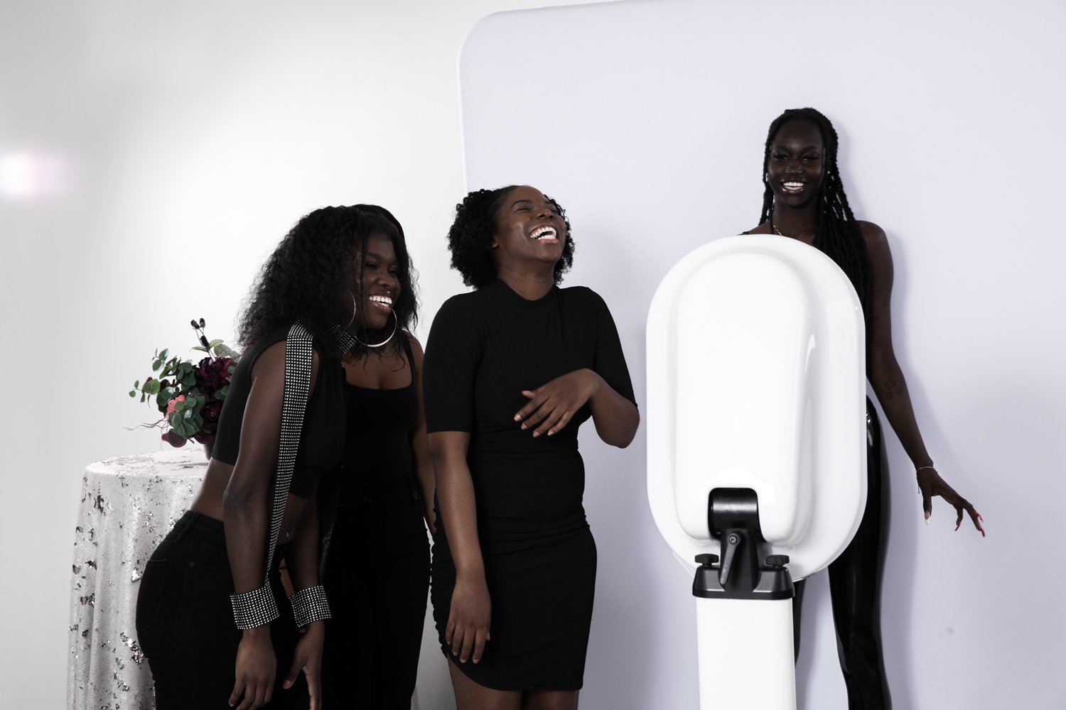 Three women laughing and smiling, standing next to a white photo booth against a white wall.