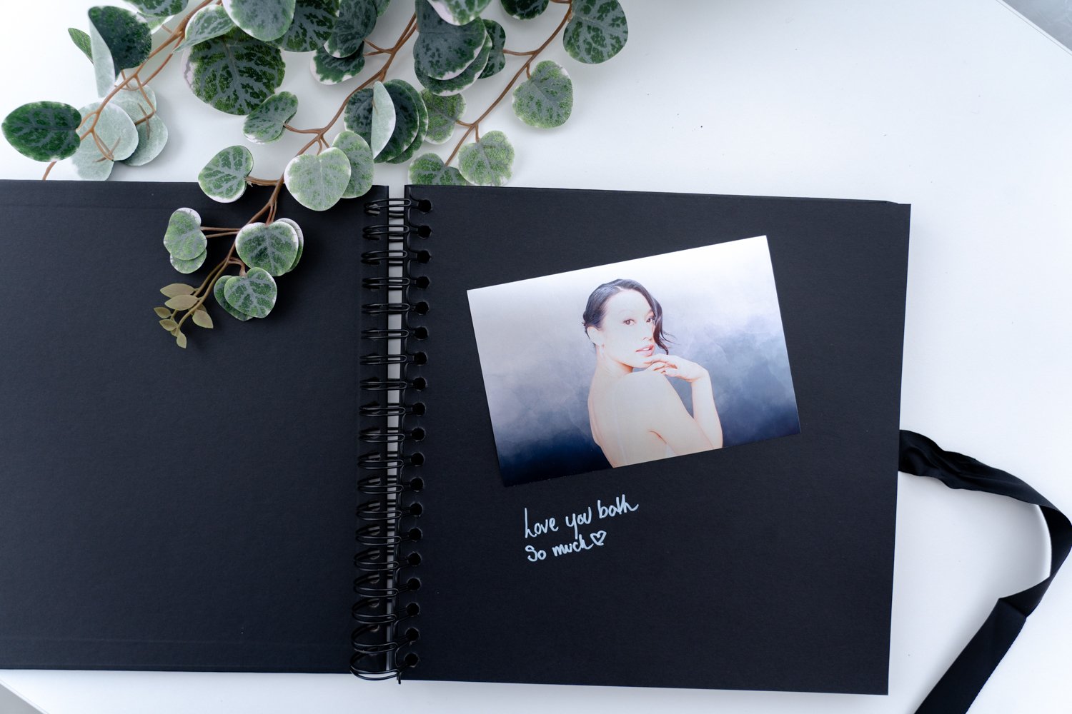 Open black photo album with a portrait of a woman and handwritten note that says, 'love you both so much' with a heart, on a white surface next to green leafy plant.