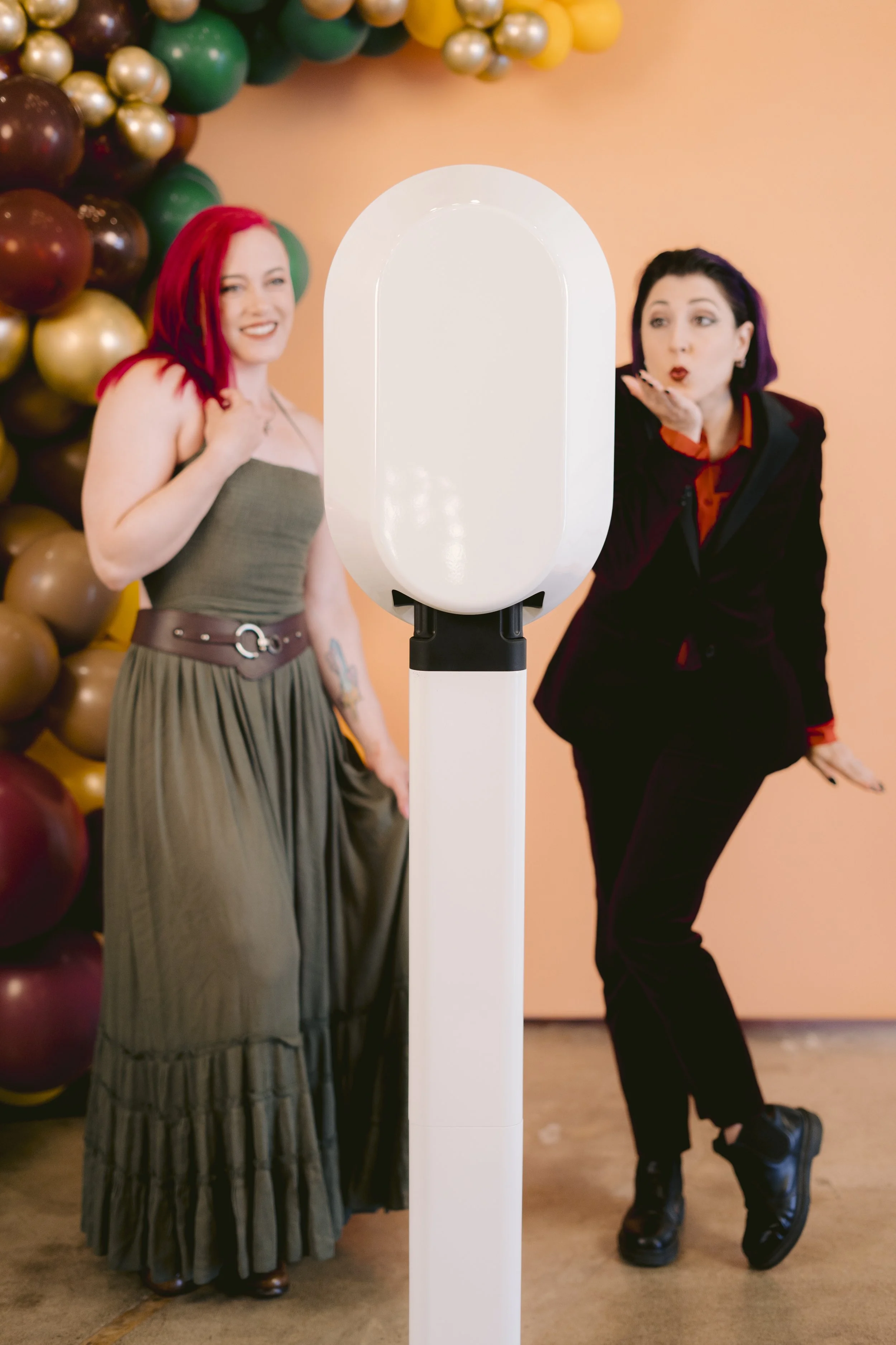 Two women standing behind a large white photo booth with a peach-colored wall and balloons in the background, one woman is blowing a kiss and the other is smiling.