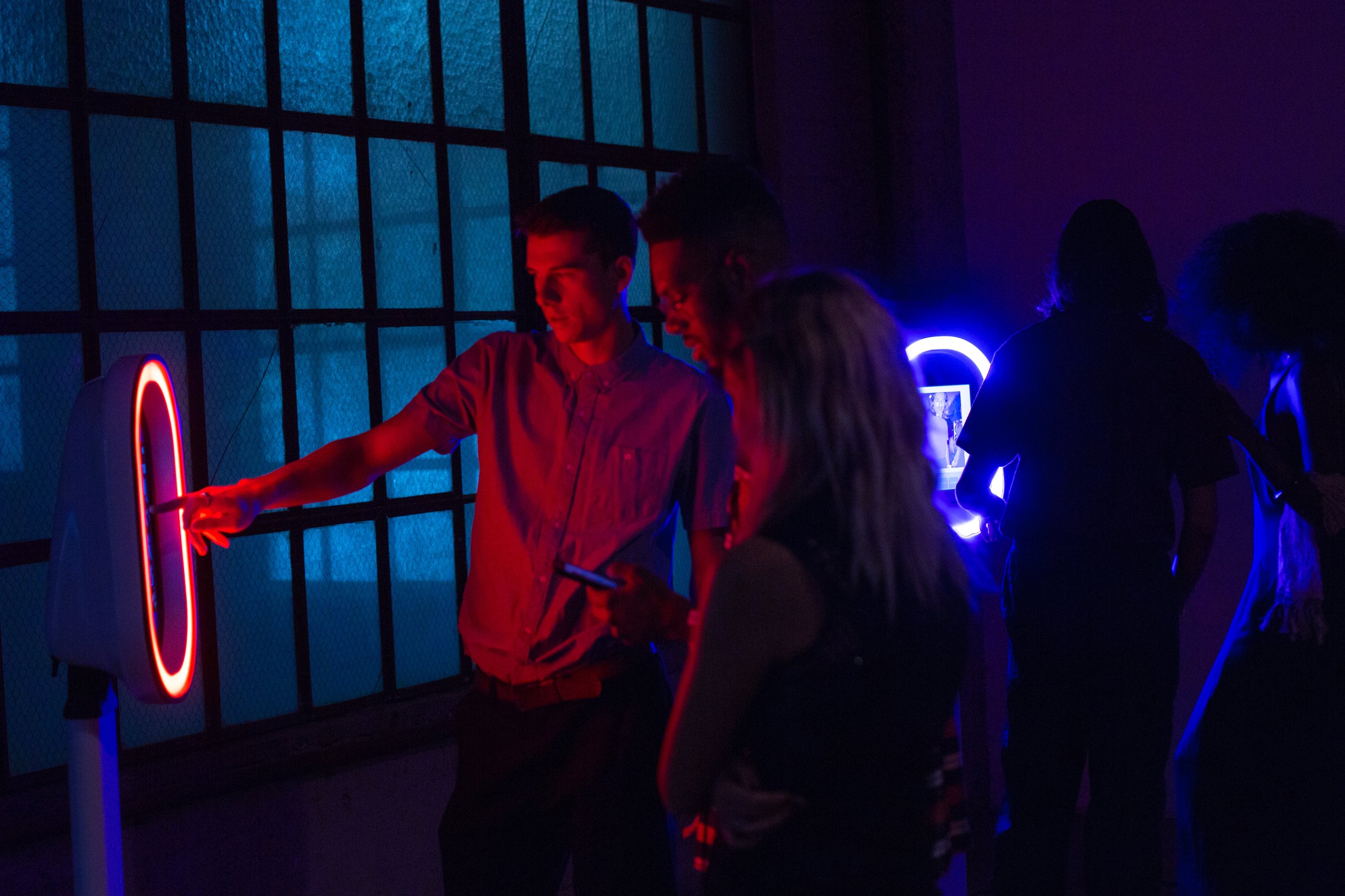 People in a dark room with neon lights, some looking at their phones, others interacting with illuminated installations, and silhouettes of individuals in the background.