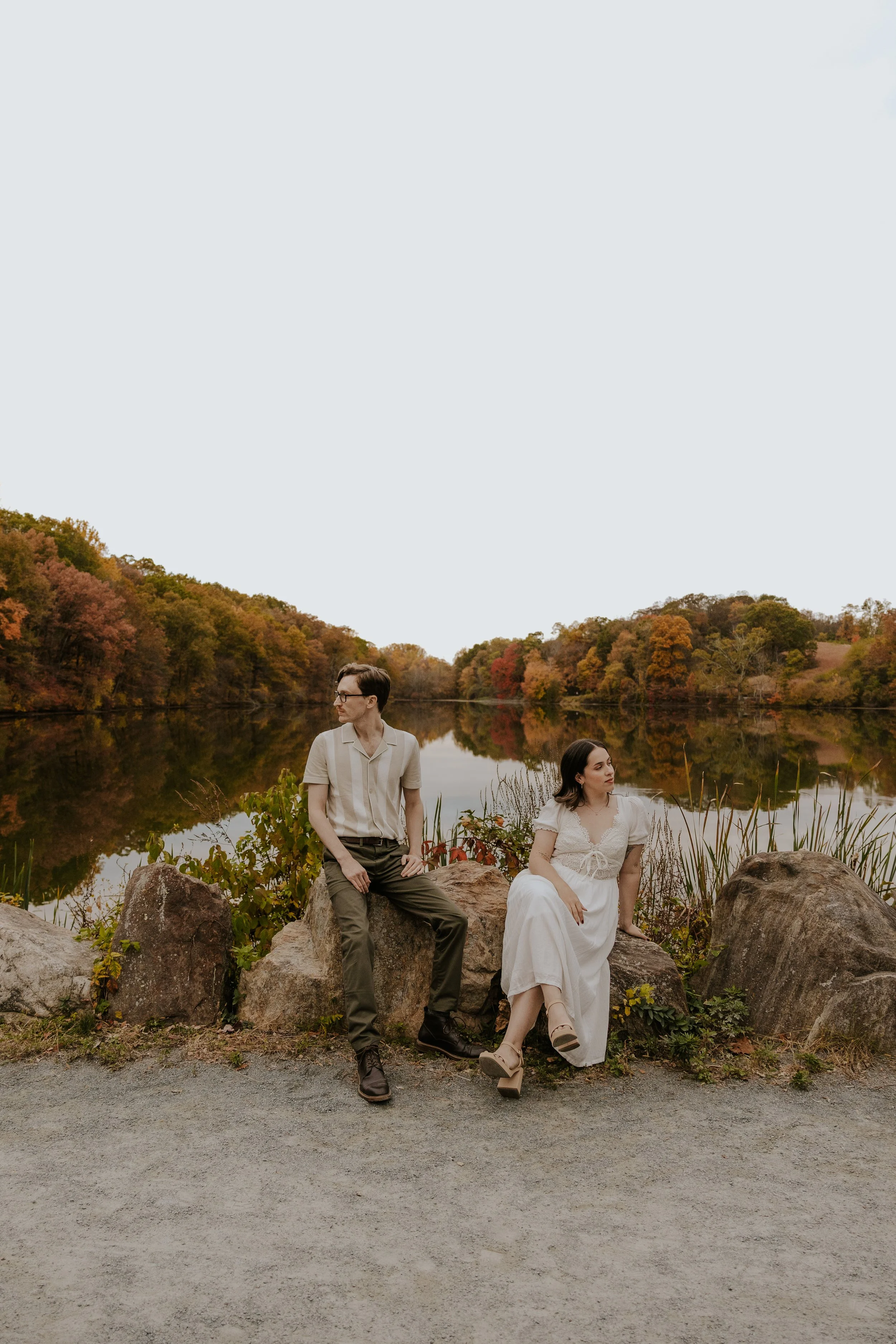 A man and woman sitting on rocks by a lake during autumn, with trees displaying fall colors reflected in the water at the Rockerfellar State Park preserve