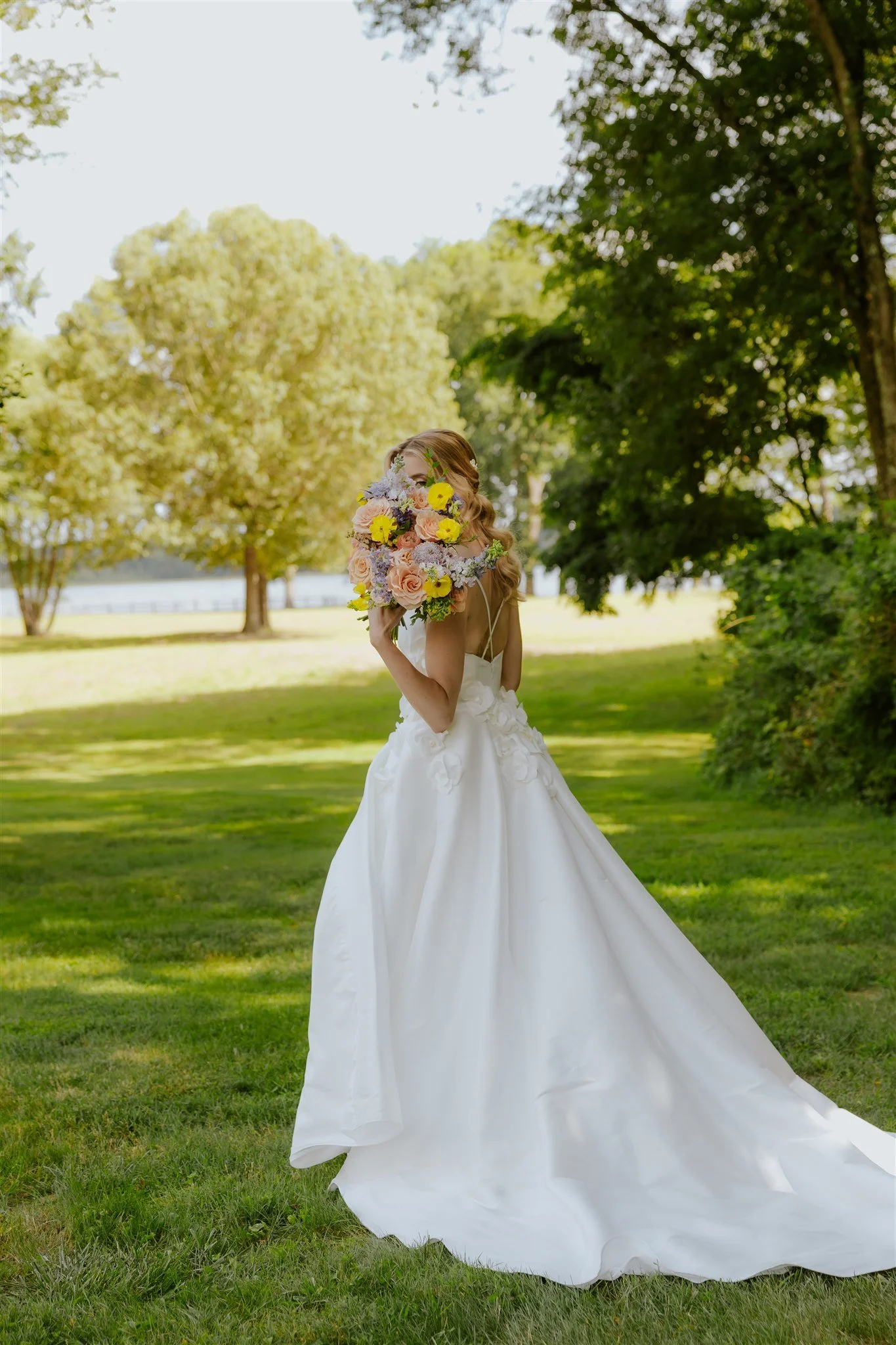 A bride standing outdoors in a green park, wearing a white wedding dress and holding a colorful bouquet of flowers in front of her face captured by a Hudson valley wedding photographer
