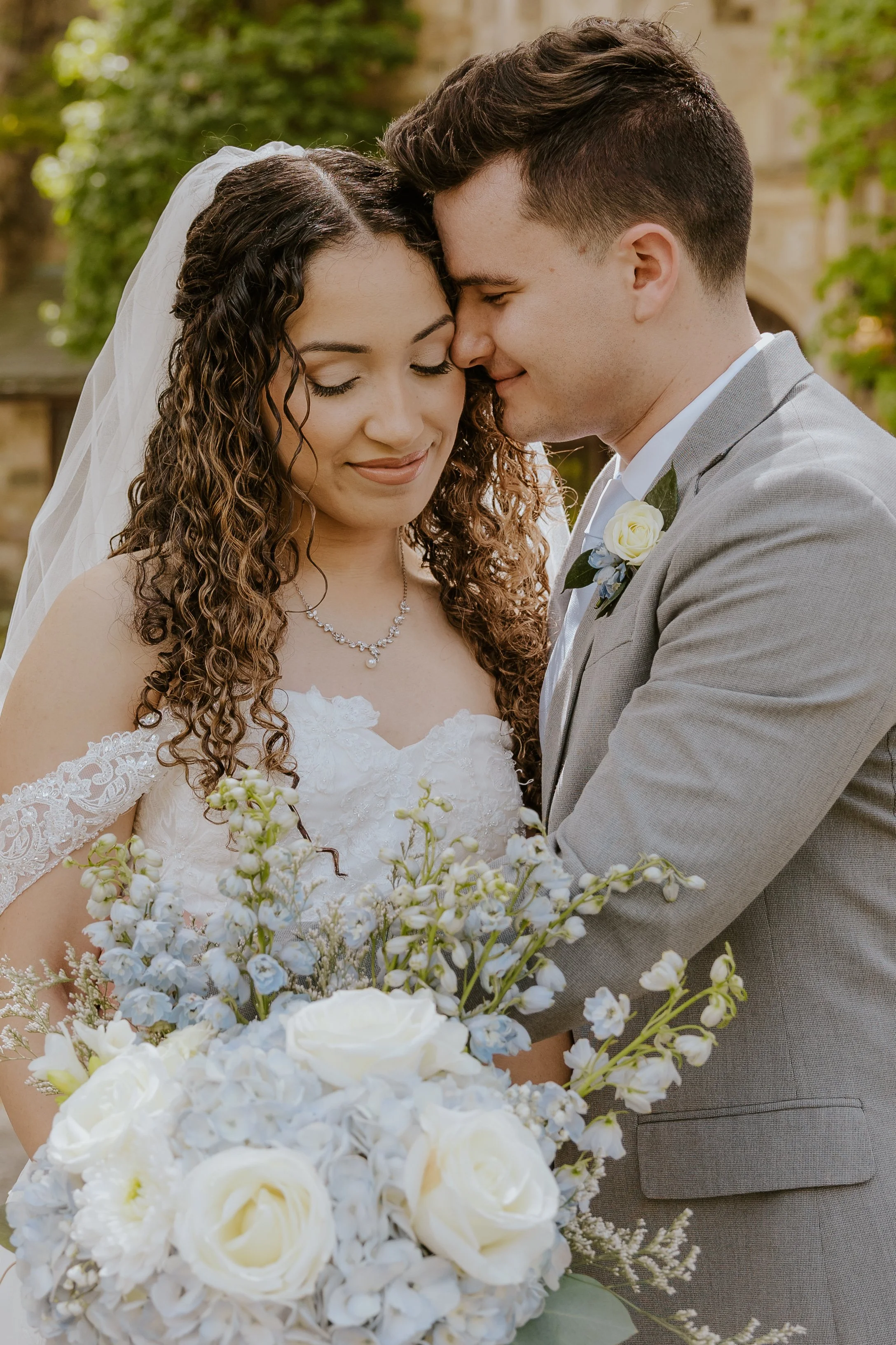 Bride and groom with foreheads touching, eyes closed, holding a bouquet of white and blue flowers, outdoors on a wedding day at the New Jersey Botanical Garden 