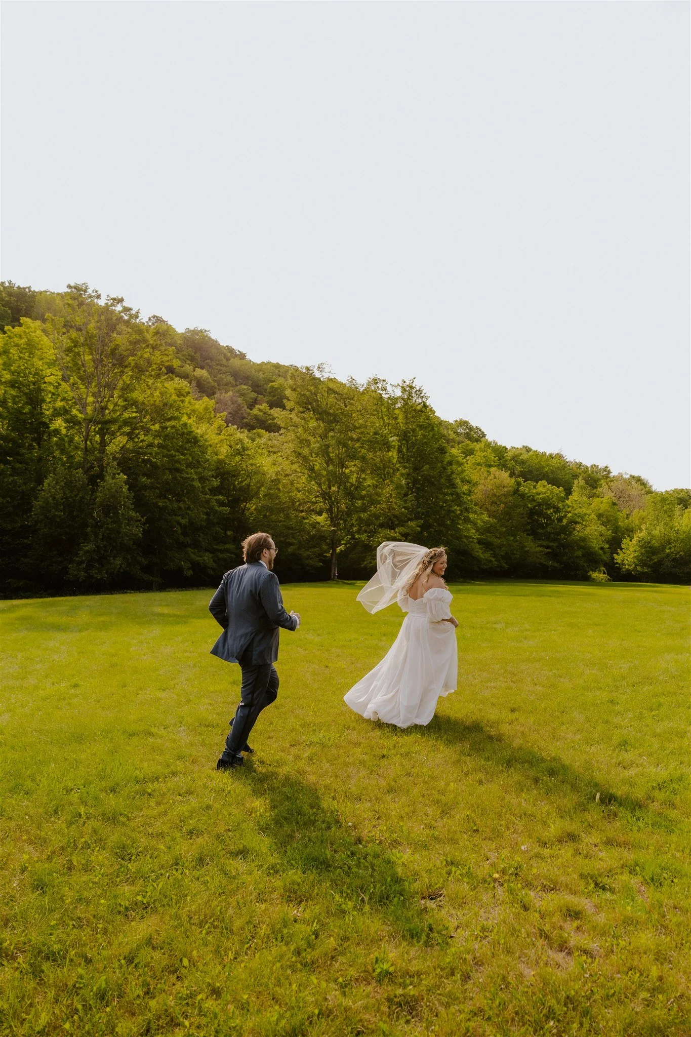 A bride and groom running on a grassy field with a forested hill in the background during daytime at Full moon resort in the Catskills on New York