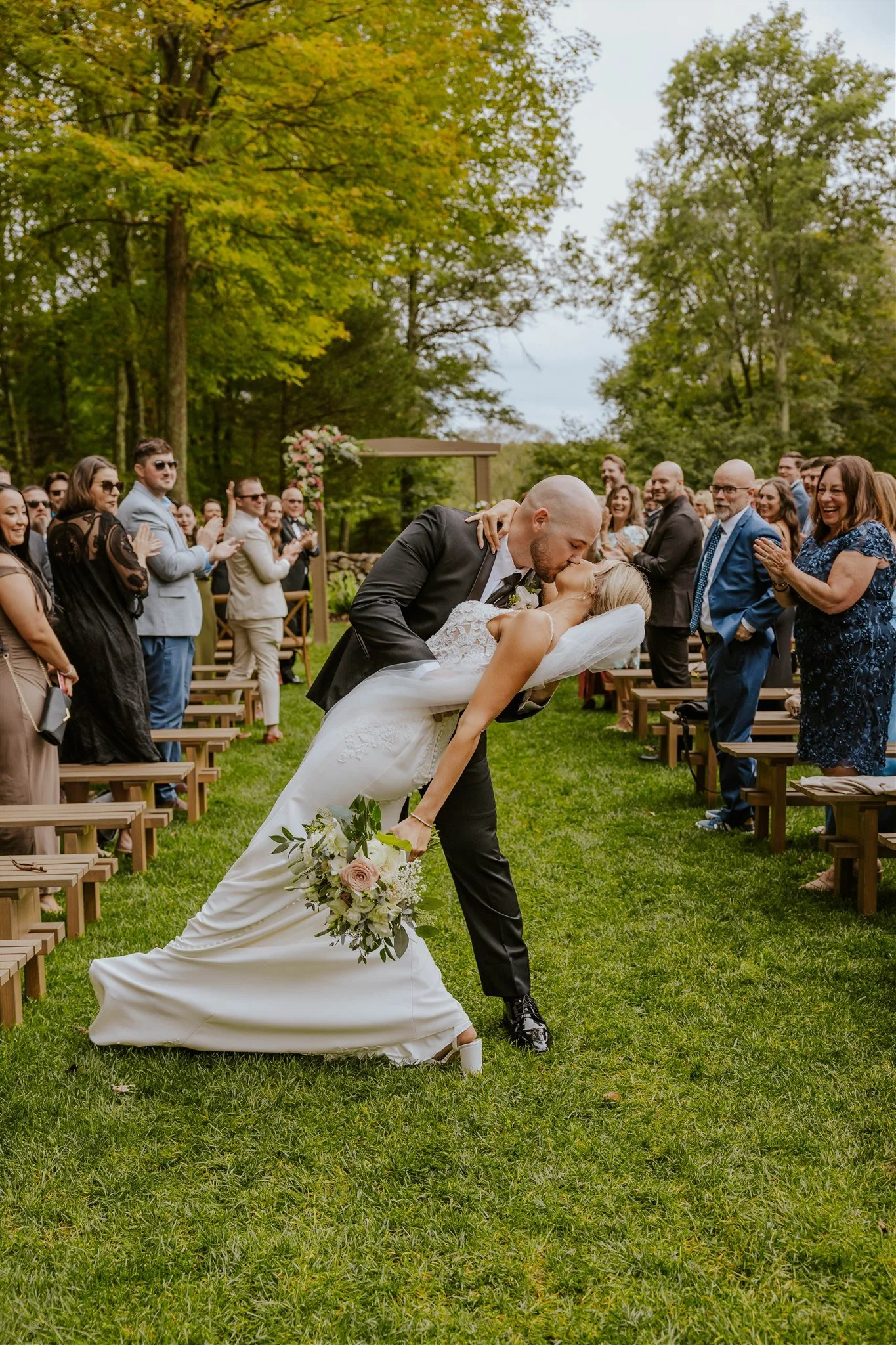 A bride and groom kissing during their outdoor wedding ceremony with guests clapping and smiling in the background in the winding hills venue in Montgomery New york