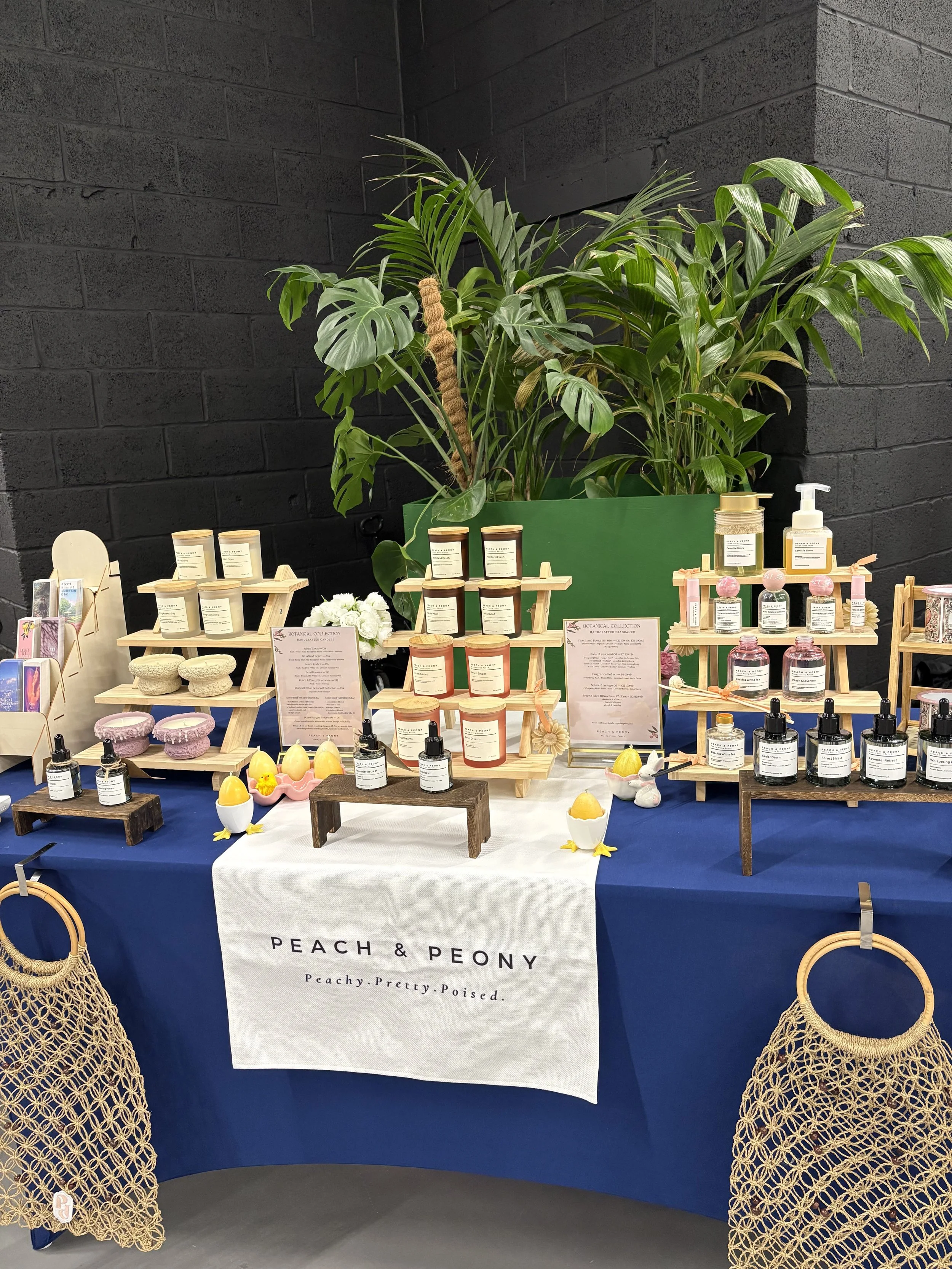 Display of natural products including candles, sprays, and bath bombs at a market stall with a large indoor plant in the background. The stall has a white banner with the brand name 'PEACH & PEONY' and the slogan 'Peachy. Pretty. Poised.' The setup i