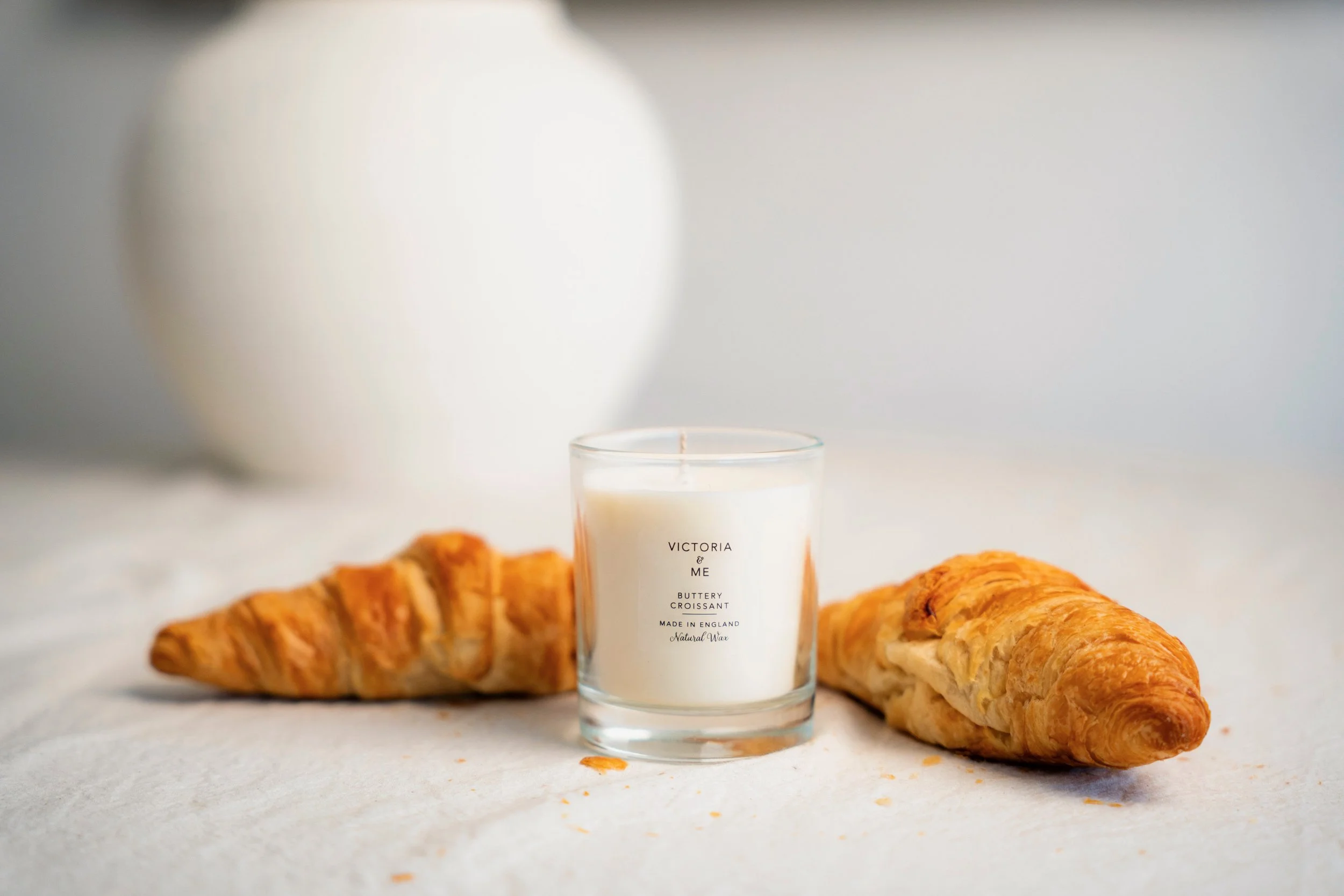 A glass candle with the words 'Victoria & Me', 'Buttery Croissant', 'Made in England', 'Natural Wax' on it, flanked by two croissants on a white surface, with a blurred background and a white candle in the background.