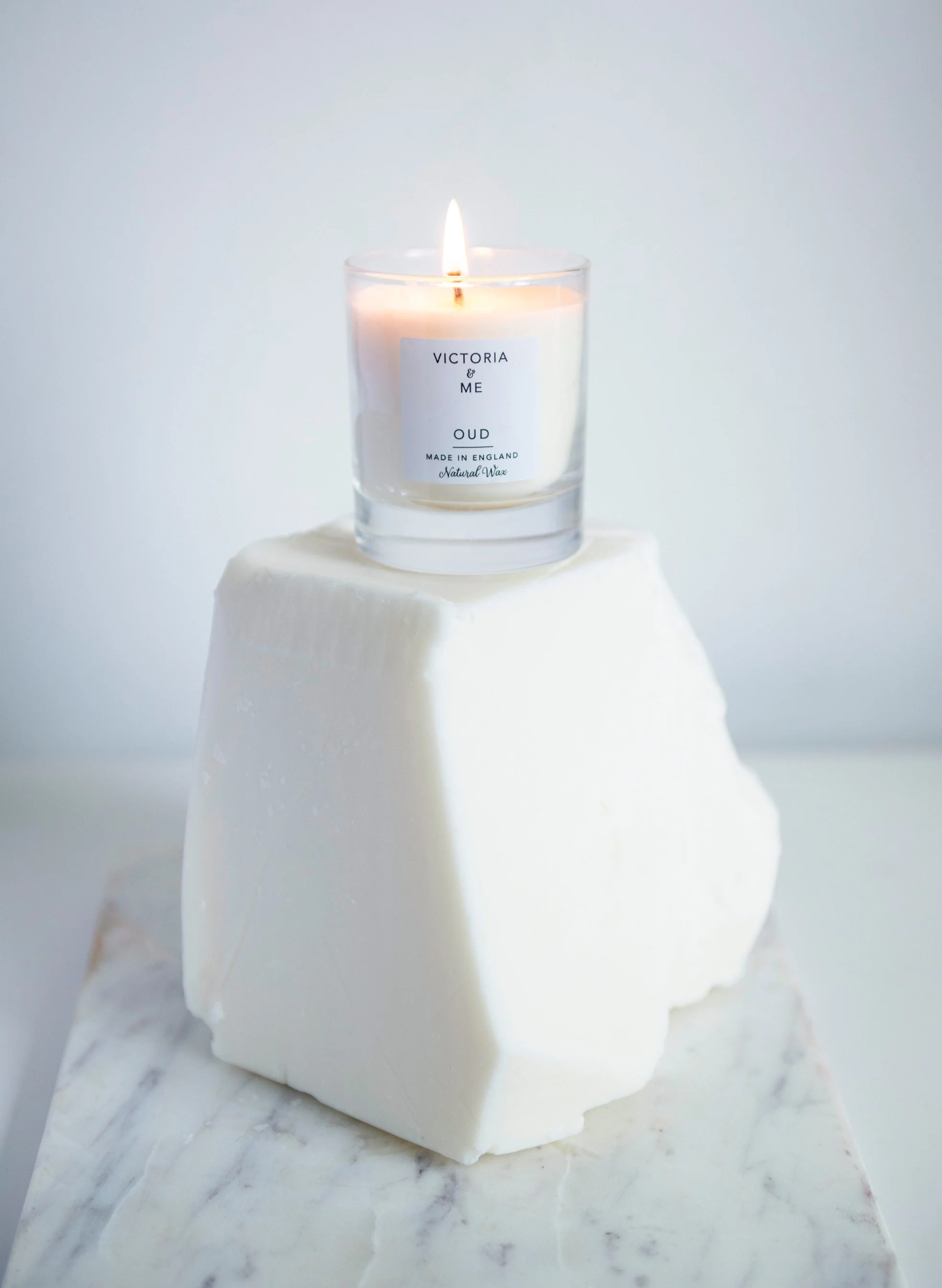 White candle labeled 'Victoria & Me Oud Made in England Natural Wax' burning in a clear glass holder, placed on a white marble surface.