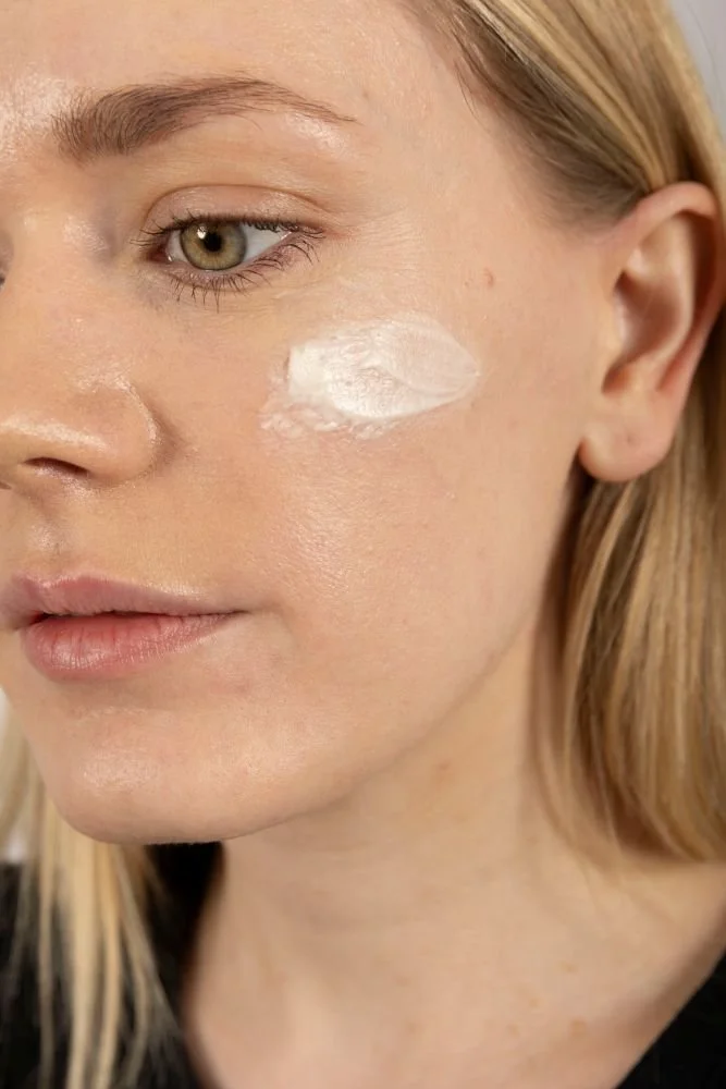 A woman with blonde hair applies white cream from Dewy Skin to her cheek.